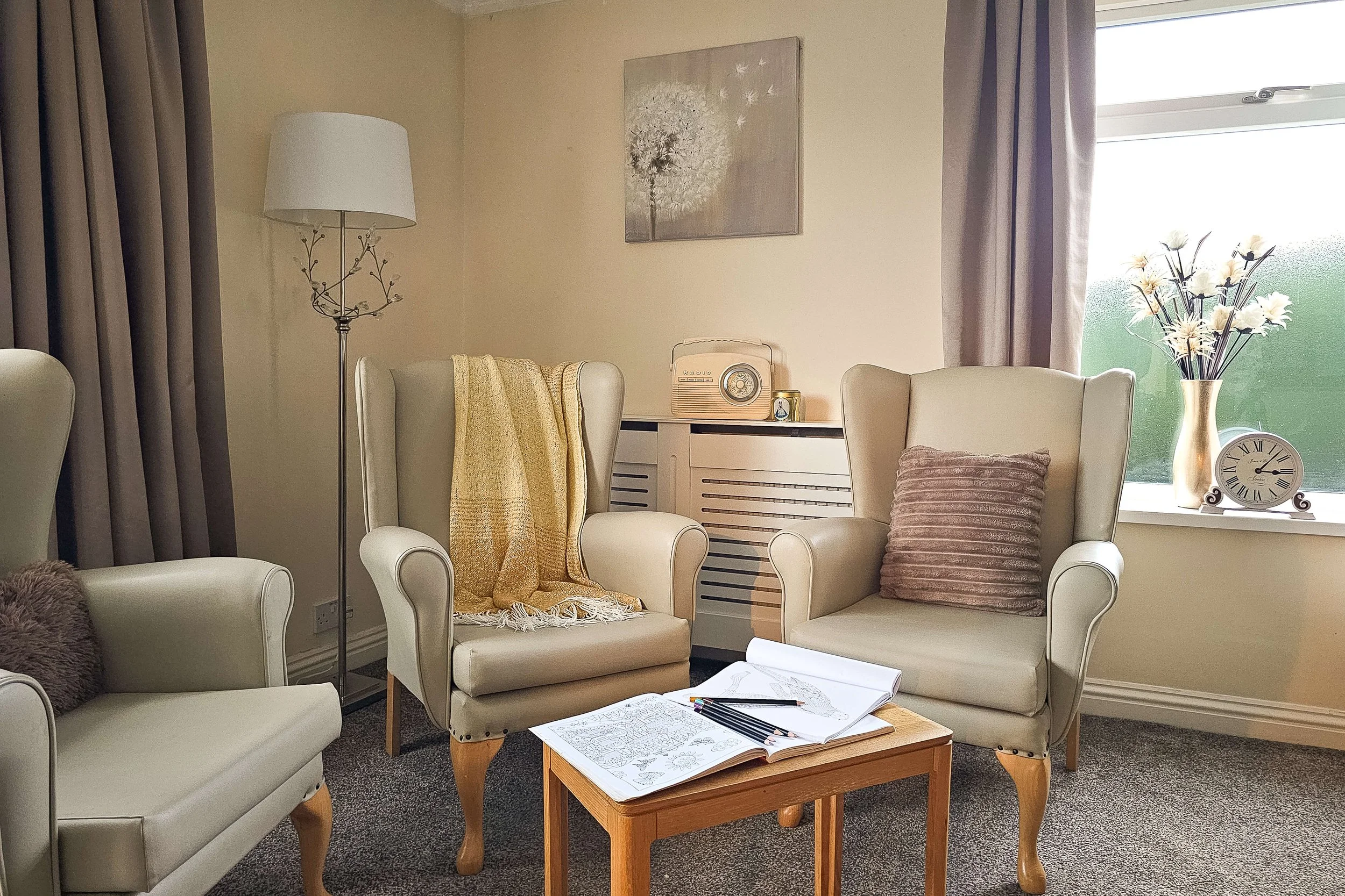 A cozy living room with two beige wingback chairs, one with a brown cushion and a yellow throw, a small wooden table with open coloring books and colored pencils, a white armchair, a tall floor lamp with a white shade, a painting of a dandelion on th