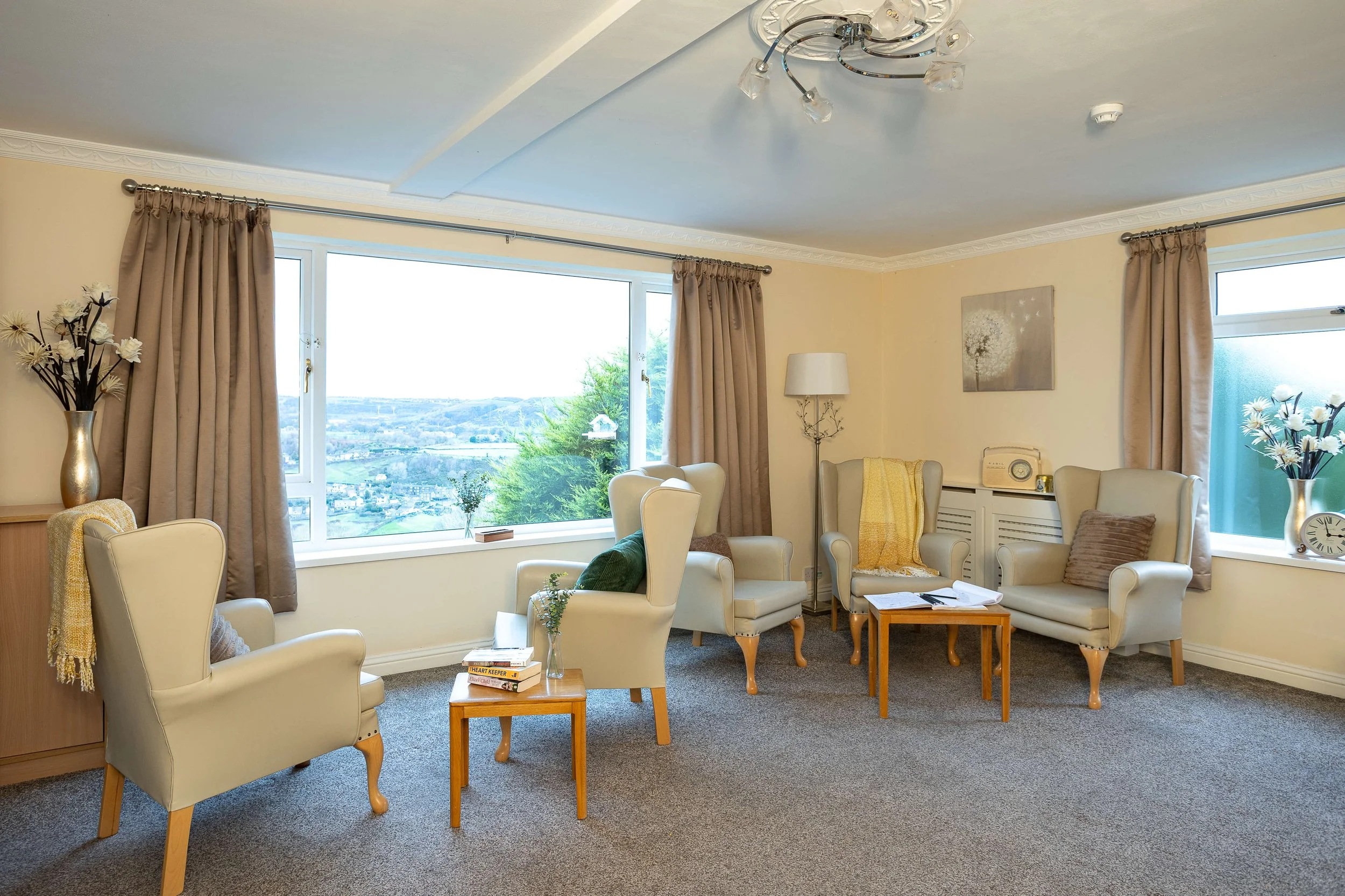 A cozy living room with four cream-colored armchairs with pillows, small wooden side tables, a floor lamp, a window with beige curtains showing a view of trees and hills, and decorative vases with flowers. There is also a painting on the wall, a vint