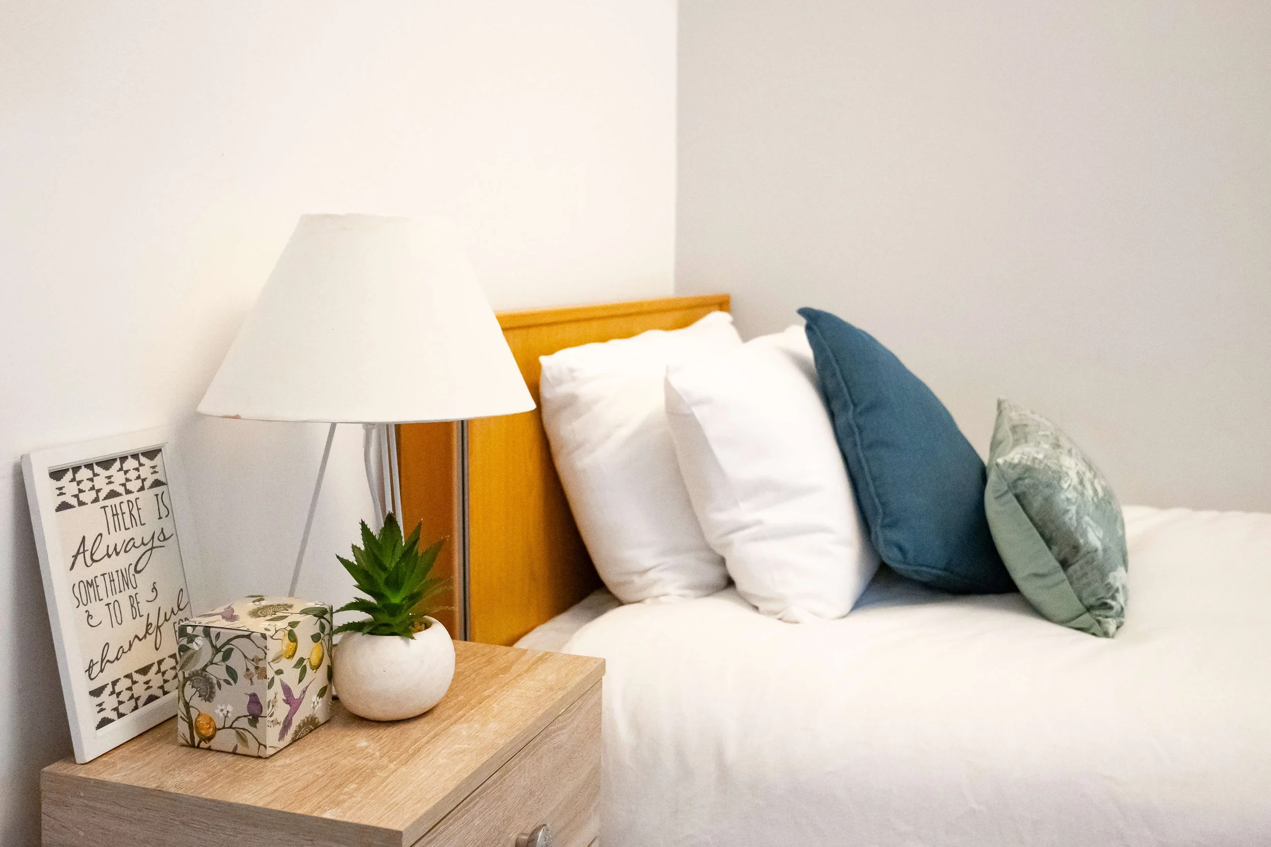 A bedroom with a bed, white pillows, and decorative pillows, a nightstand with a lamp, a framed quote, a plant, and a decorative box