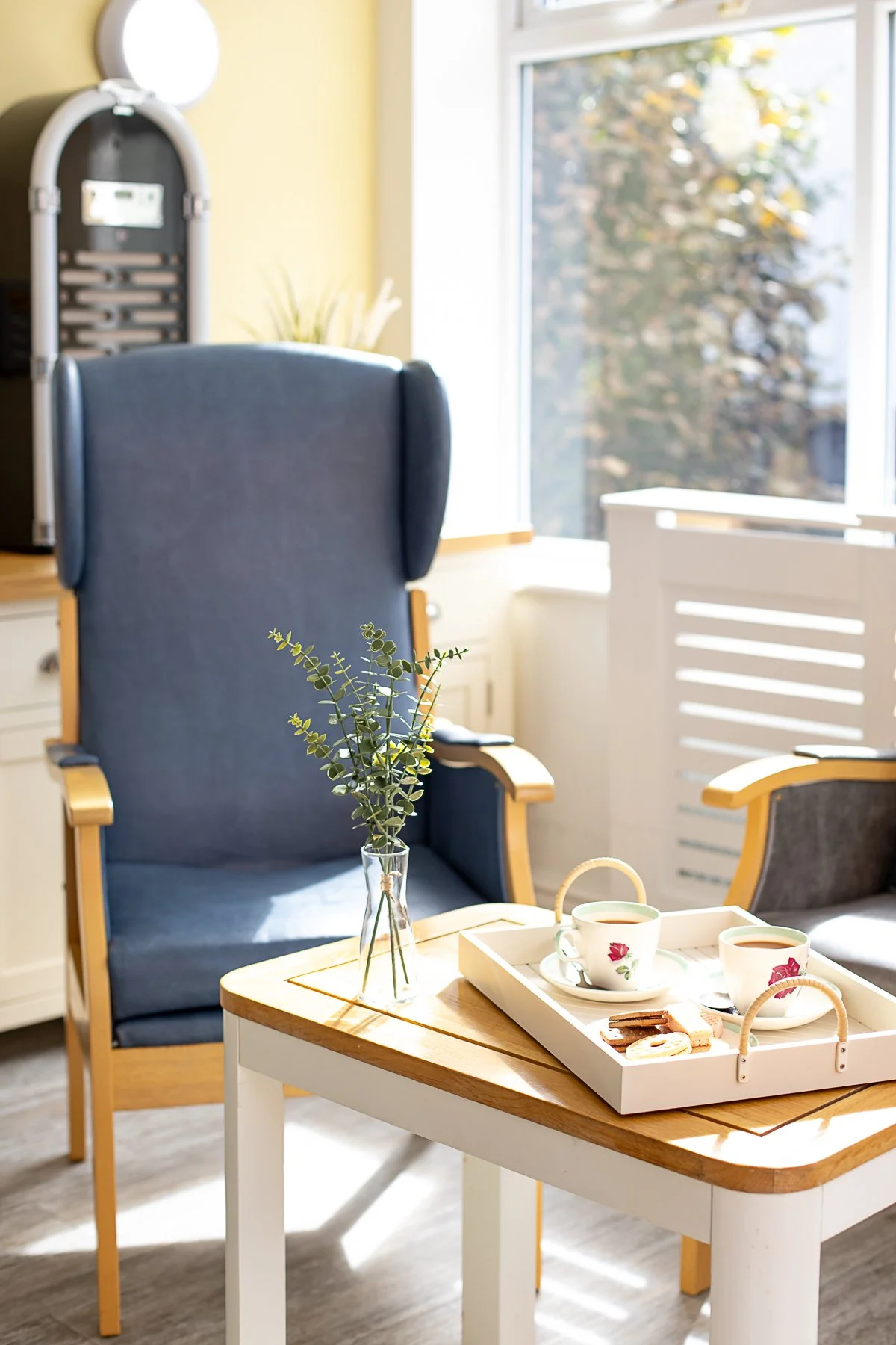 A cozy sunlit corner of a room with two chairs, a small white table, a tray with tea cups and snacks, a small vase with green foliage, and a large window showing outdoor trees.