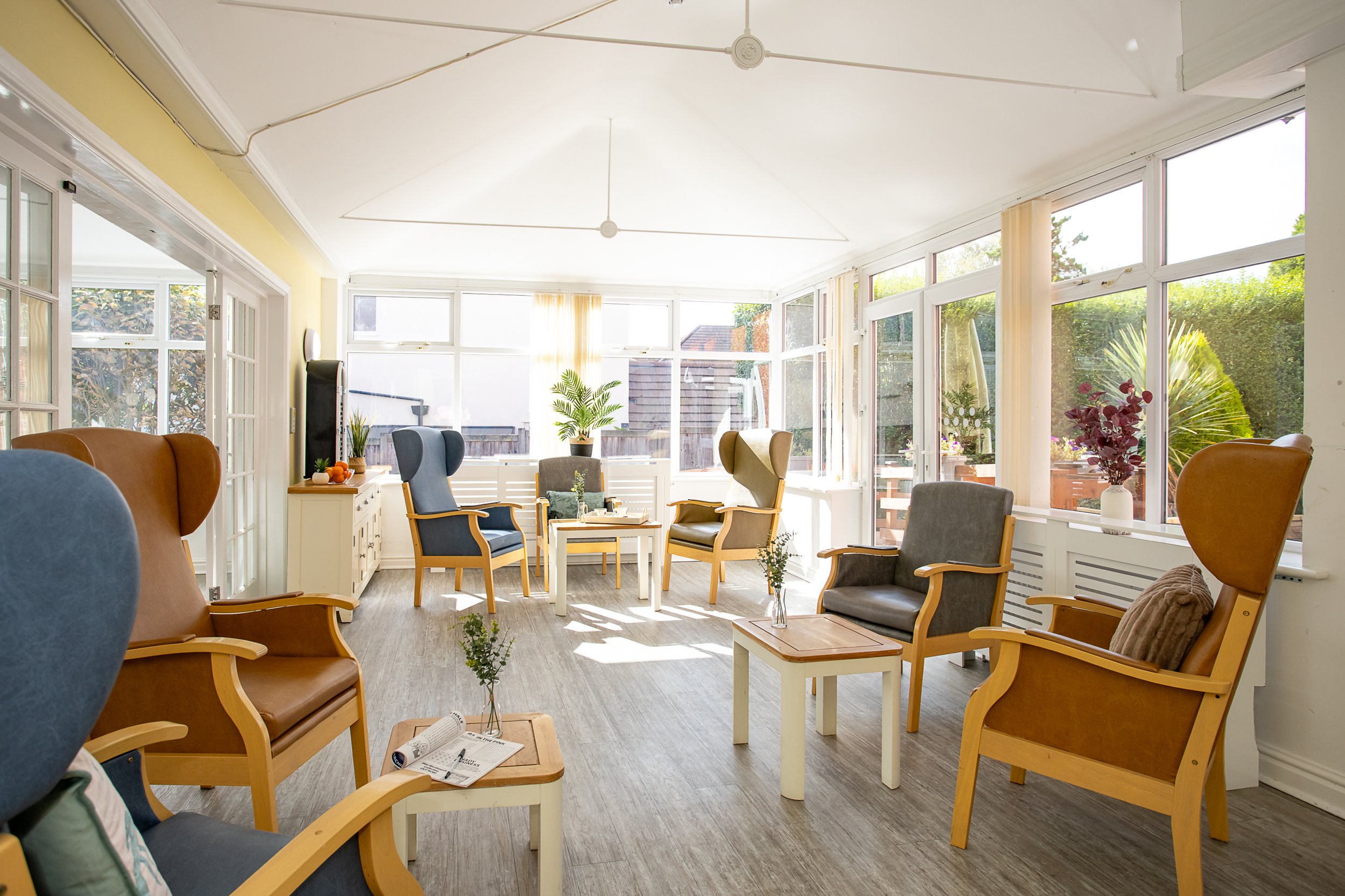 Bright sunlit lounge with large windows, wooden armchairs with cushioned seats and pillows, small tables with vases, potted plants, and a standing heater.