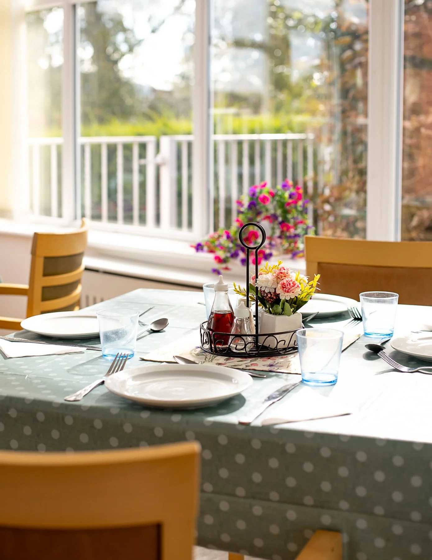 A dining table set for a meal with white plates, silverware, glasses, and a centerpiece with flowers and condiments, in front of a large window with a view of a porch and trees outside.