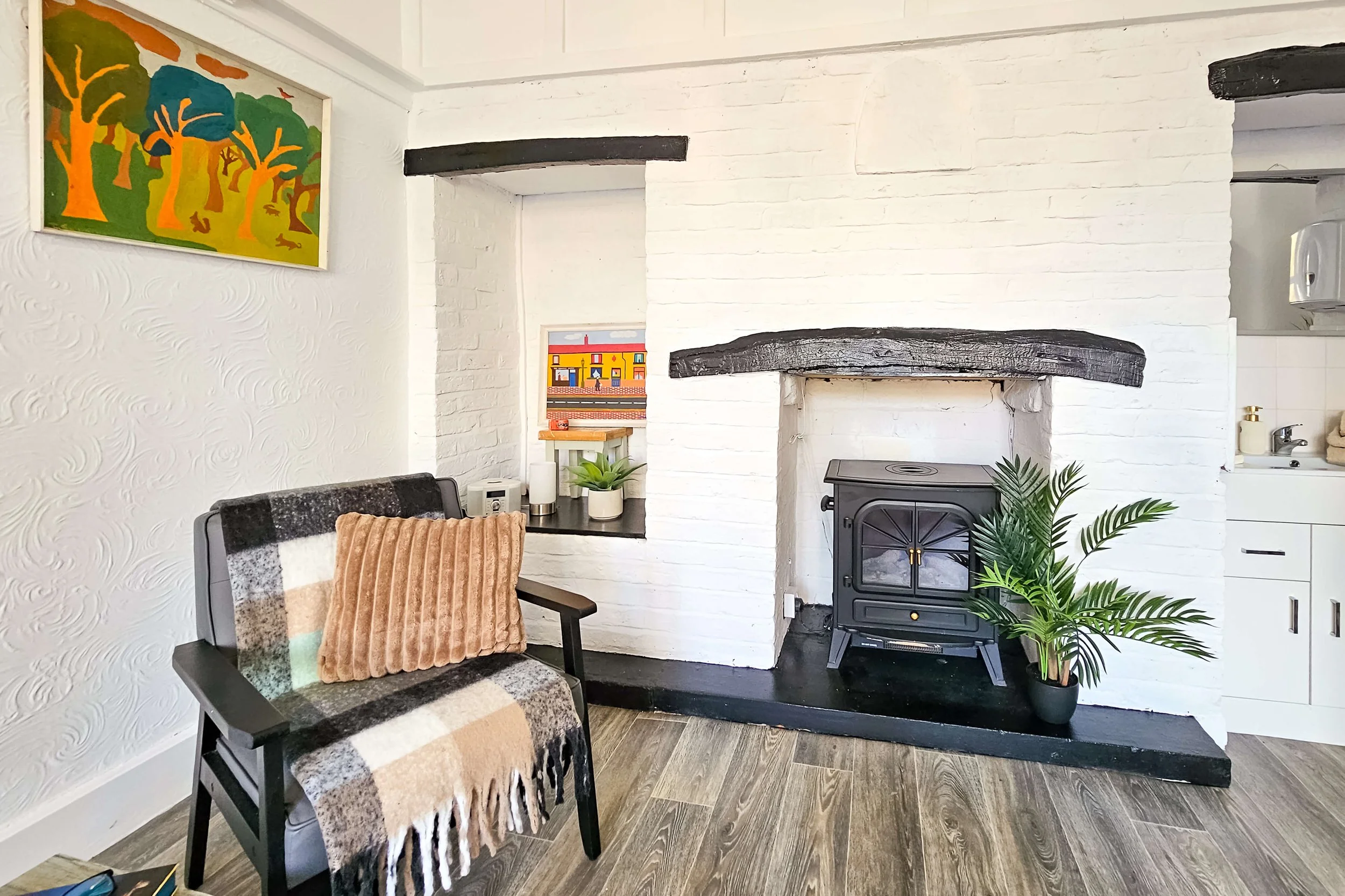 Living room corner with a white brick fireplace, black wooden beam mantel, and a black wood stove. There is a large potted green plant and a window seat with small plants and a colorful picture. A patterned blanket and a pink ribbed pillow are on a b