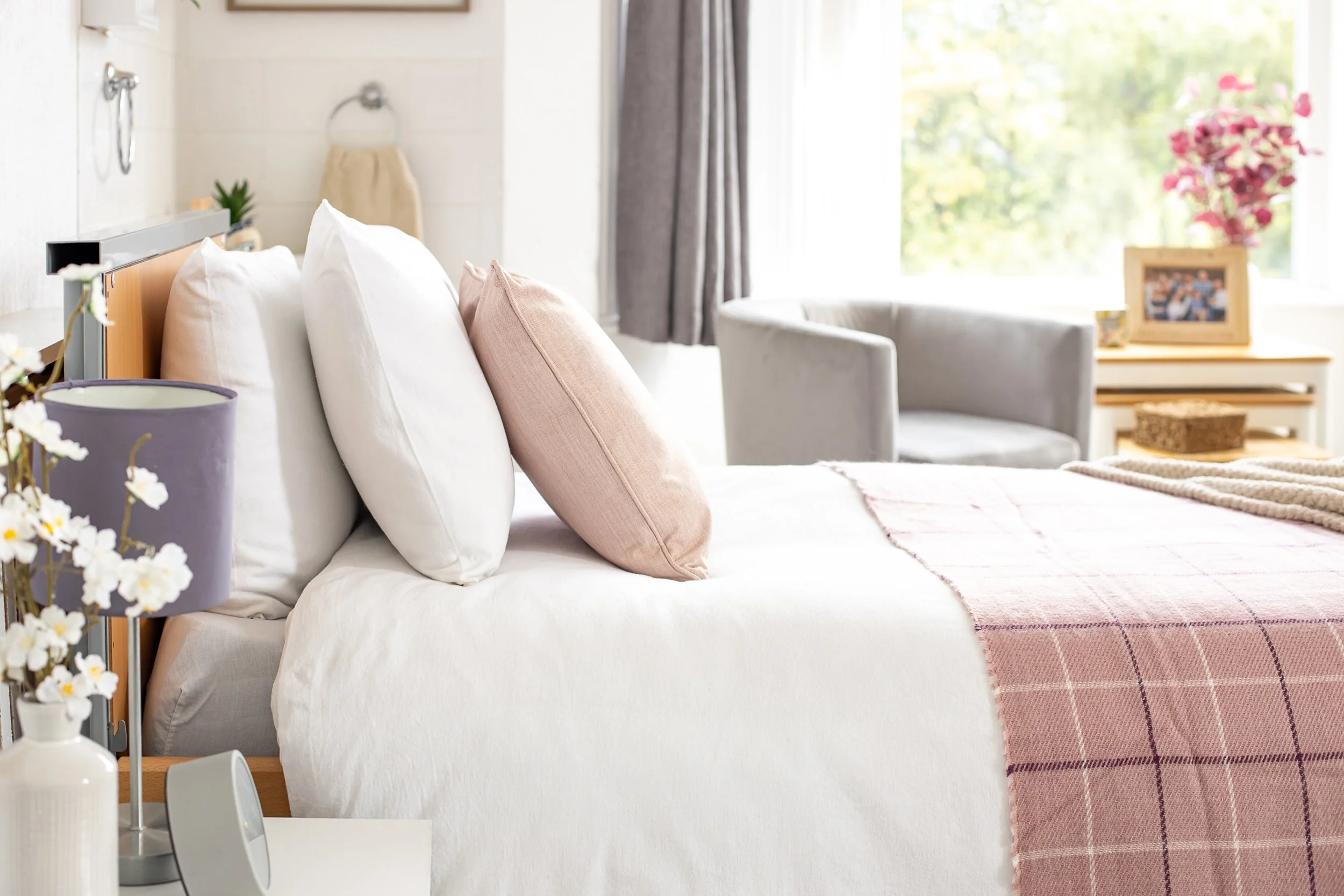 A neatly made bed with white and pink pillows, a pink and white checkered blanket, in a sunlit bedroom with a nightstand, flowers on a table, and a seating area near a window.