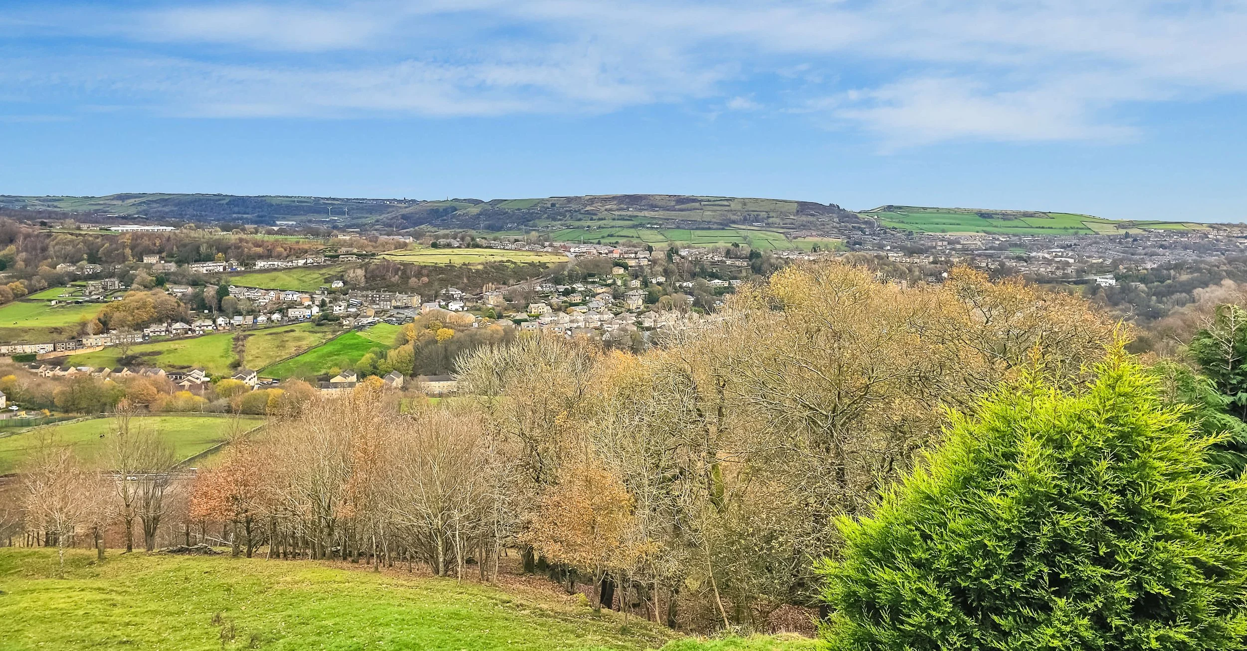 A scenic view of a countryside landscape with rolling green hills, scattered trees with some showing fall colors, and a distant town or village under a partly cloudy blue sky.