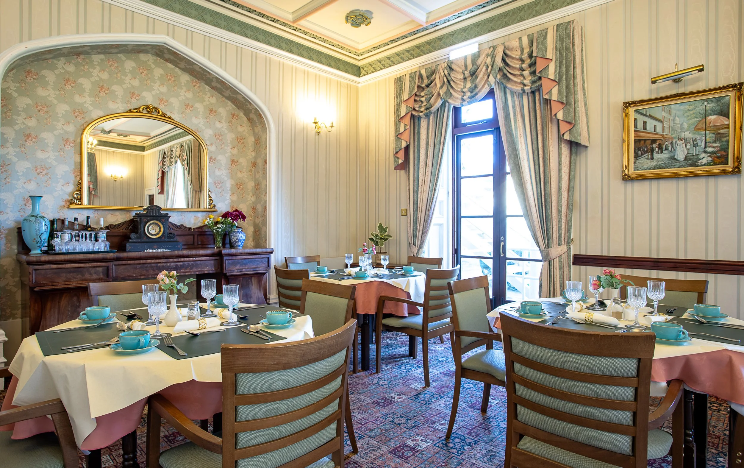 Elegant dining room with round tables set with turquoise cups, glasses, and floral centerpieces, wooden chairs, ornate mirror, vintage furniture, floral wallpaper, and heavy drapes covering glass doors leading outside.