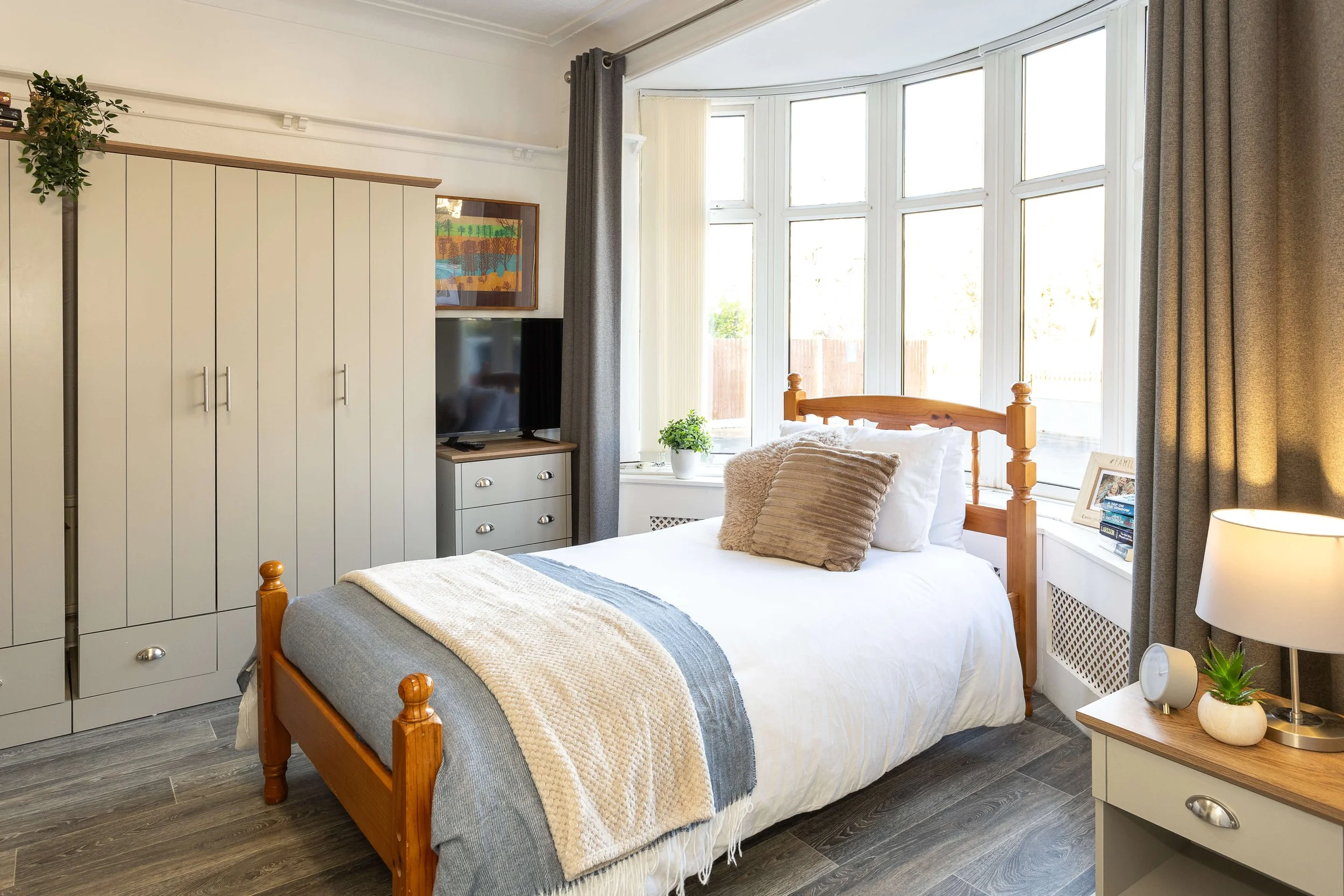 A cozy bedroom with a wooden bed, white bedding, beige and gray pillows, a side table with a lamp, a small plant, a clock, and framed photos. Large window with gray curtains allows natural light, with a wardrobe and television nearby.