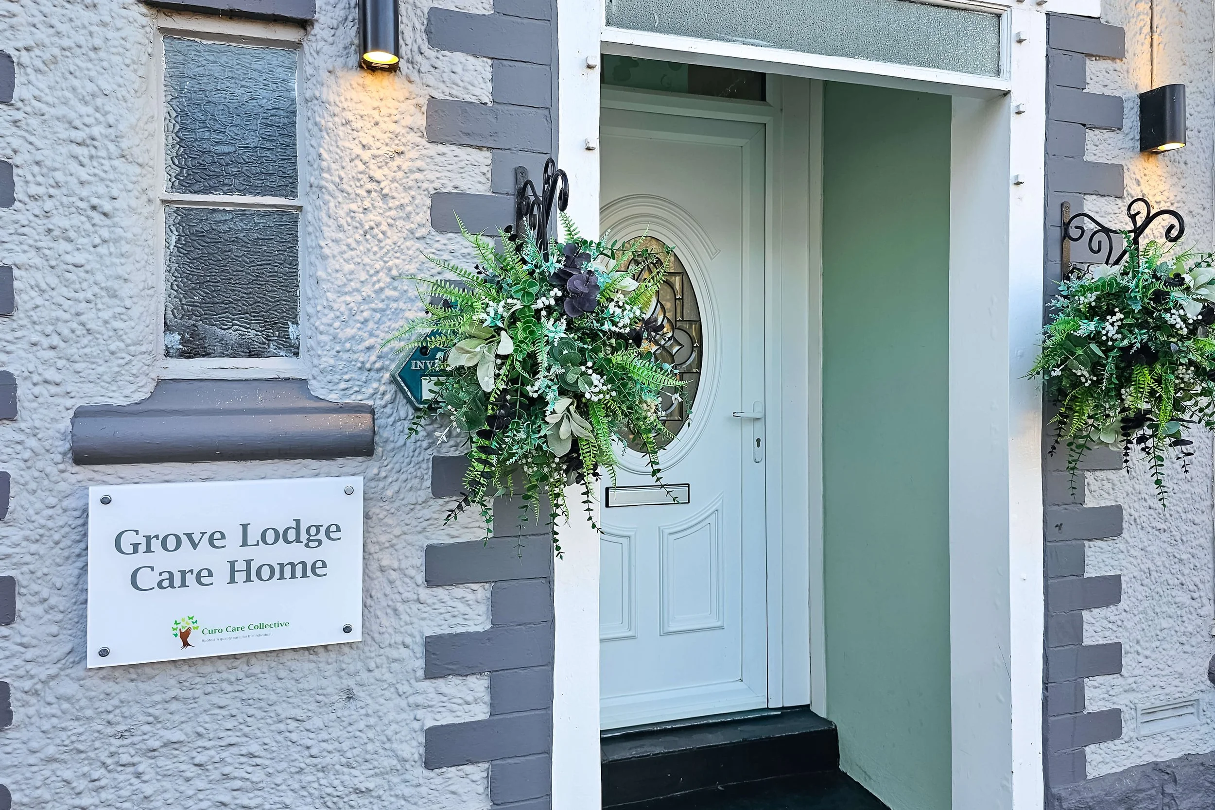 Exterior view of Grove Lodge Care Home entrance with white door, decorated with floral arrangements on both sides, and a sign that reads 'Grove Lodge Care Home'.