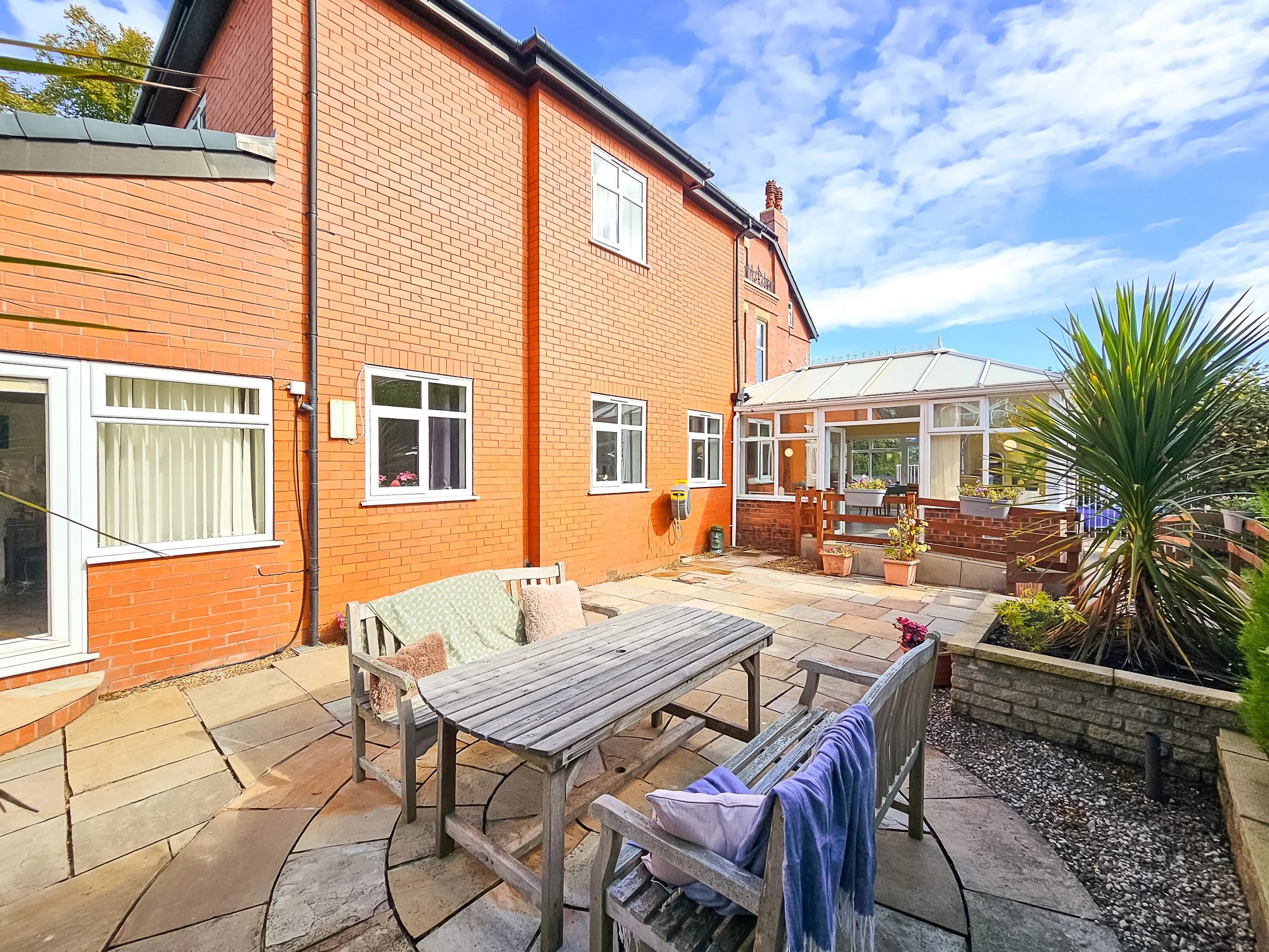 A backyard patio with wooden outdoor furniture, including a table and benches, surrounded by potted plants. The patio has flagstone paving and is adjacent to a red brick house with white-framed windows. There is a glass conservatory attached to the h