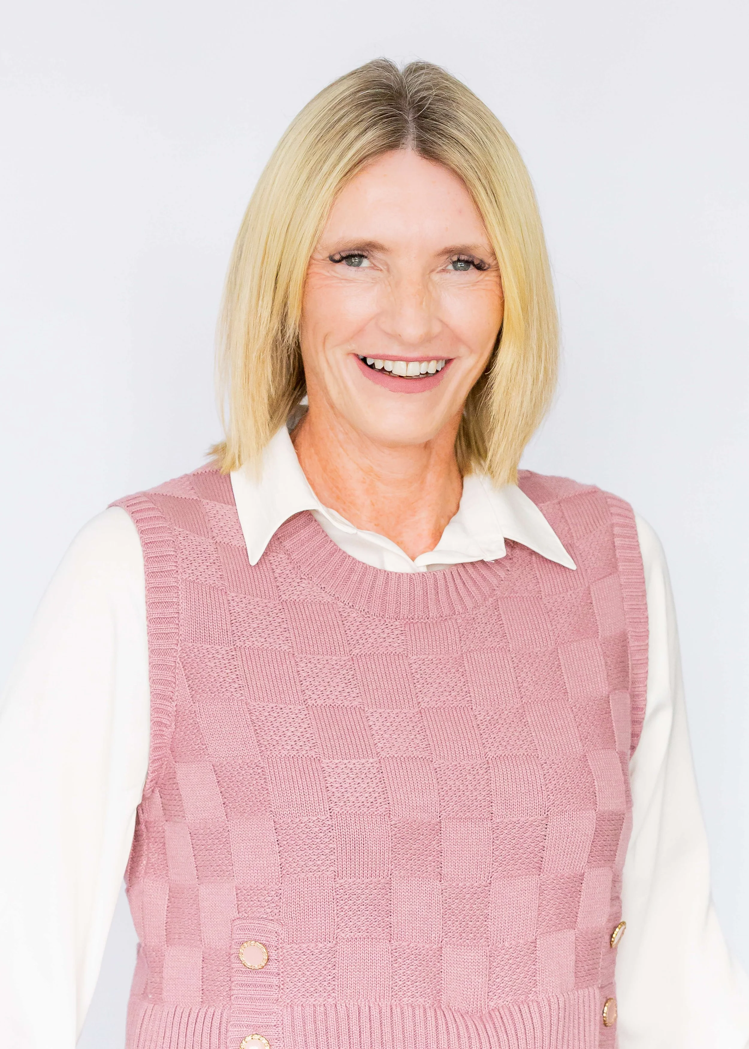 A smiling middle-aged woman with blonde hair, wearing a white collared shirt and pink knit vest, against a plain light background.