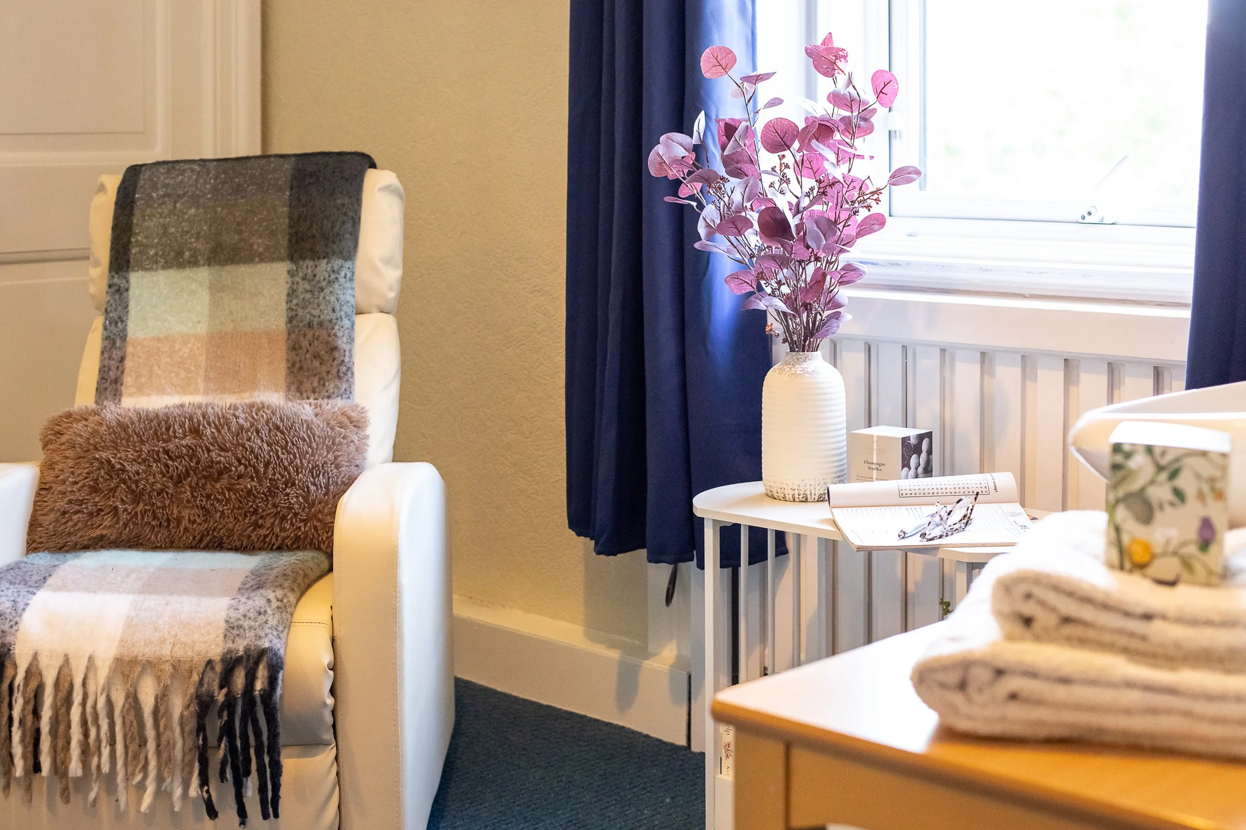 A cozy corner with a white armchair draped with a plaid blanket and a furry cushion, next to a small white table holding a vase with pink leaves, an open book, and glasses, near a window with blue curtains.