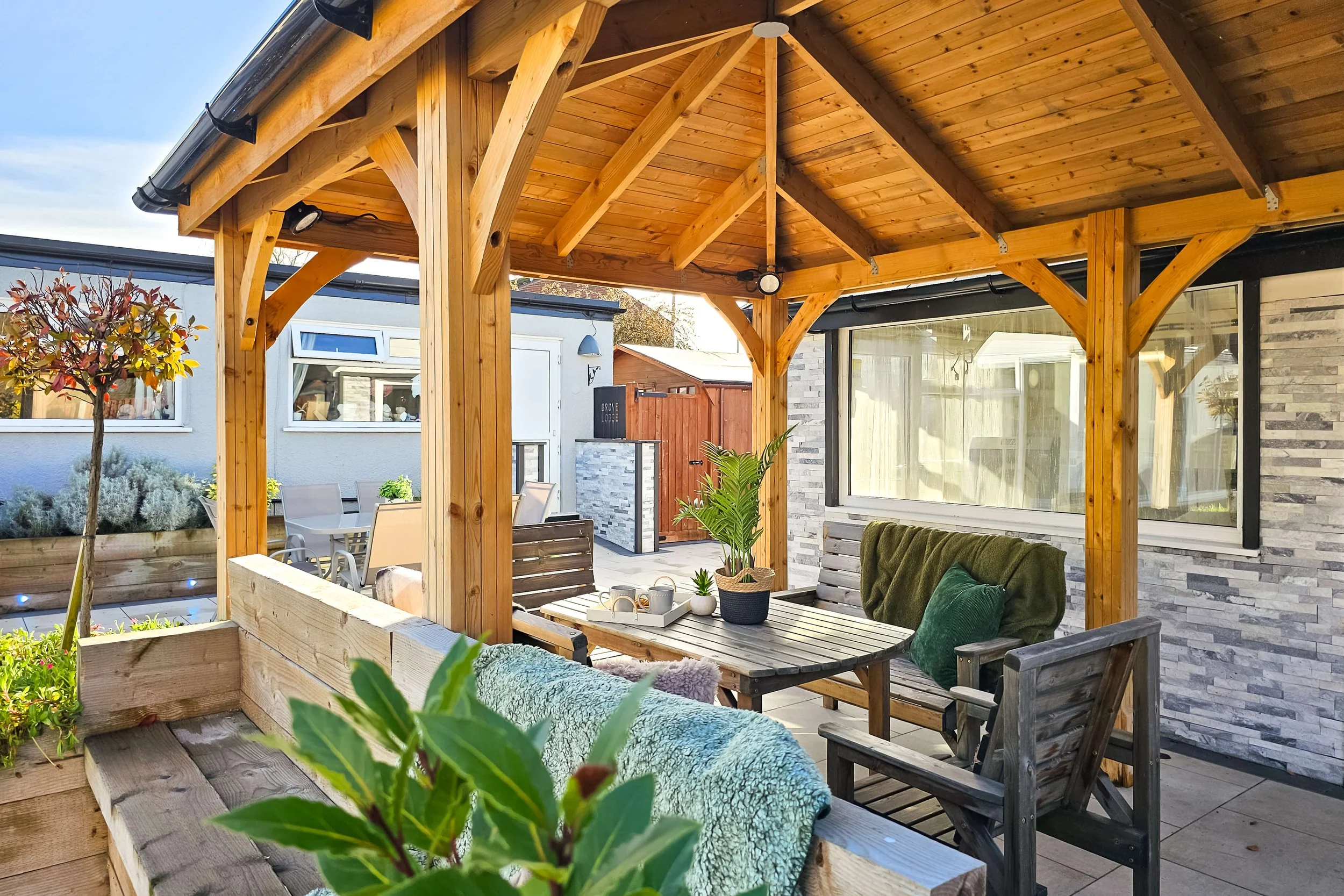 Outdoor patio with wooden gazebo, seating area with wooden chairs, a table with potted plant and cups, and a background garden with trees and plants.