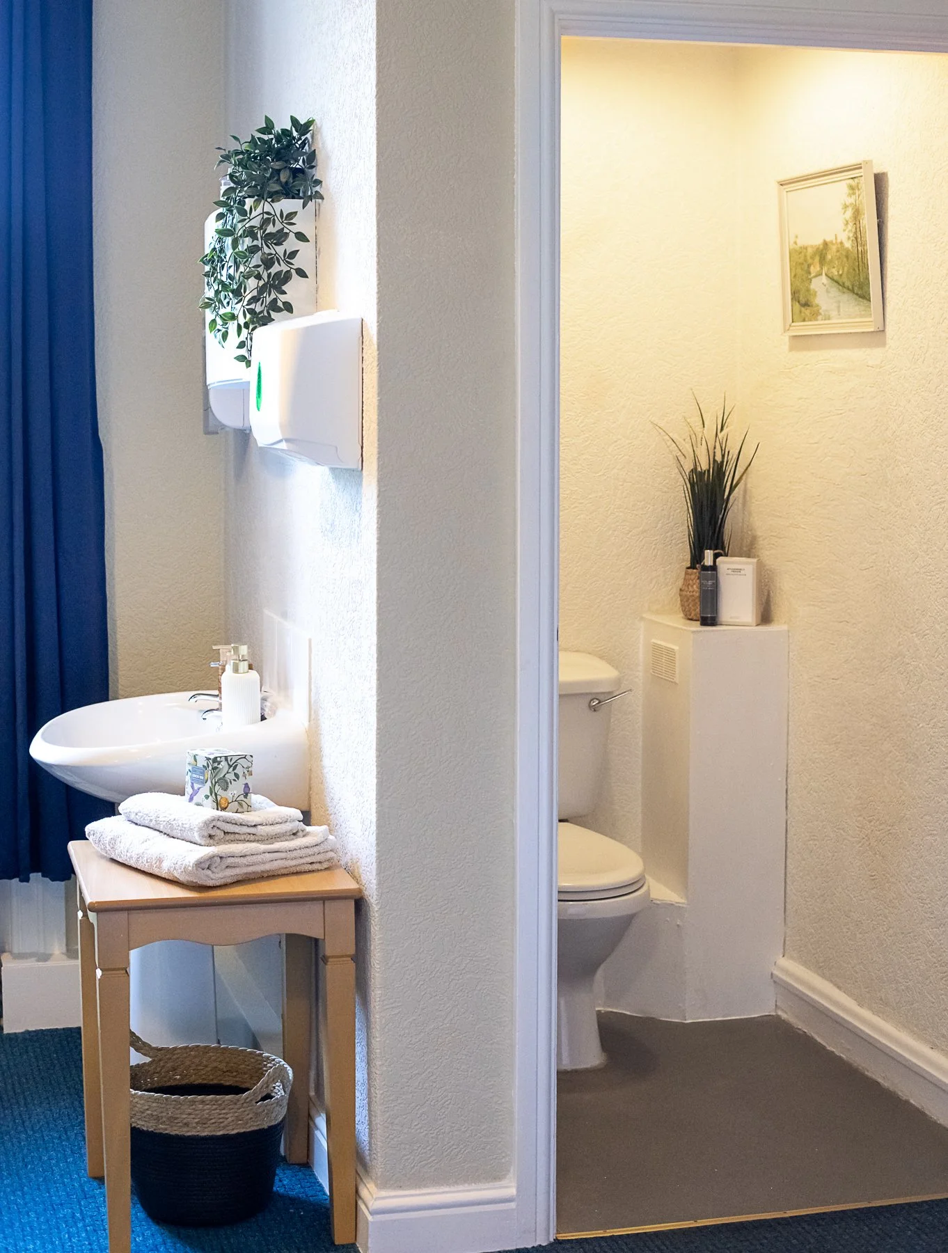 A small bathroom with a toilet and a tall potted plant, visible through a doorway. The main area has a sink with a towel and some toiletries, a small table, and a basket underneath.