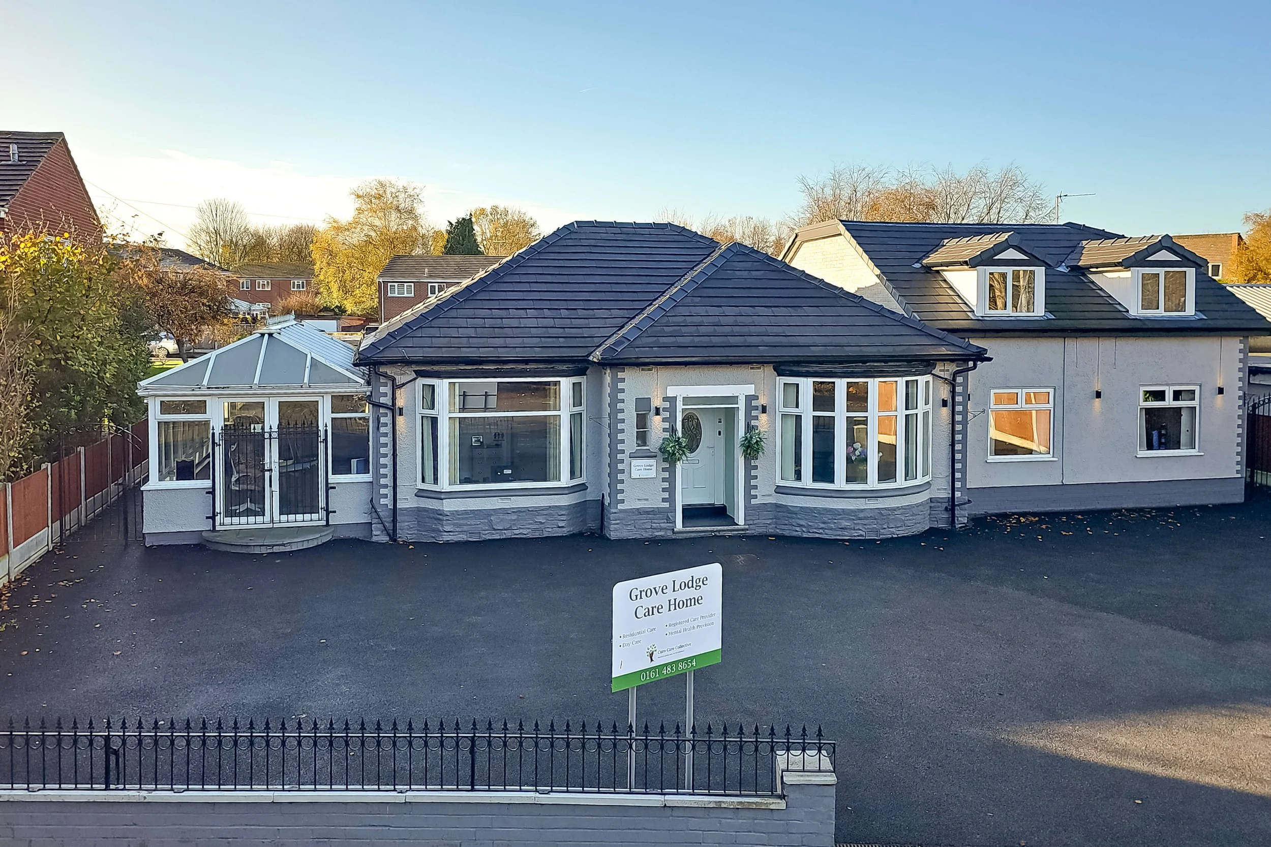 Exterior view of a care home building with a sign reading 'Grove Lodge Care Home' in front, a black driveway, and trees in the background.