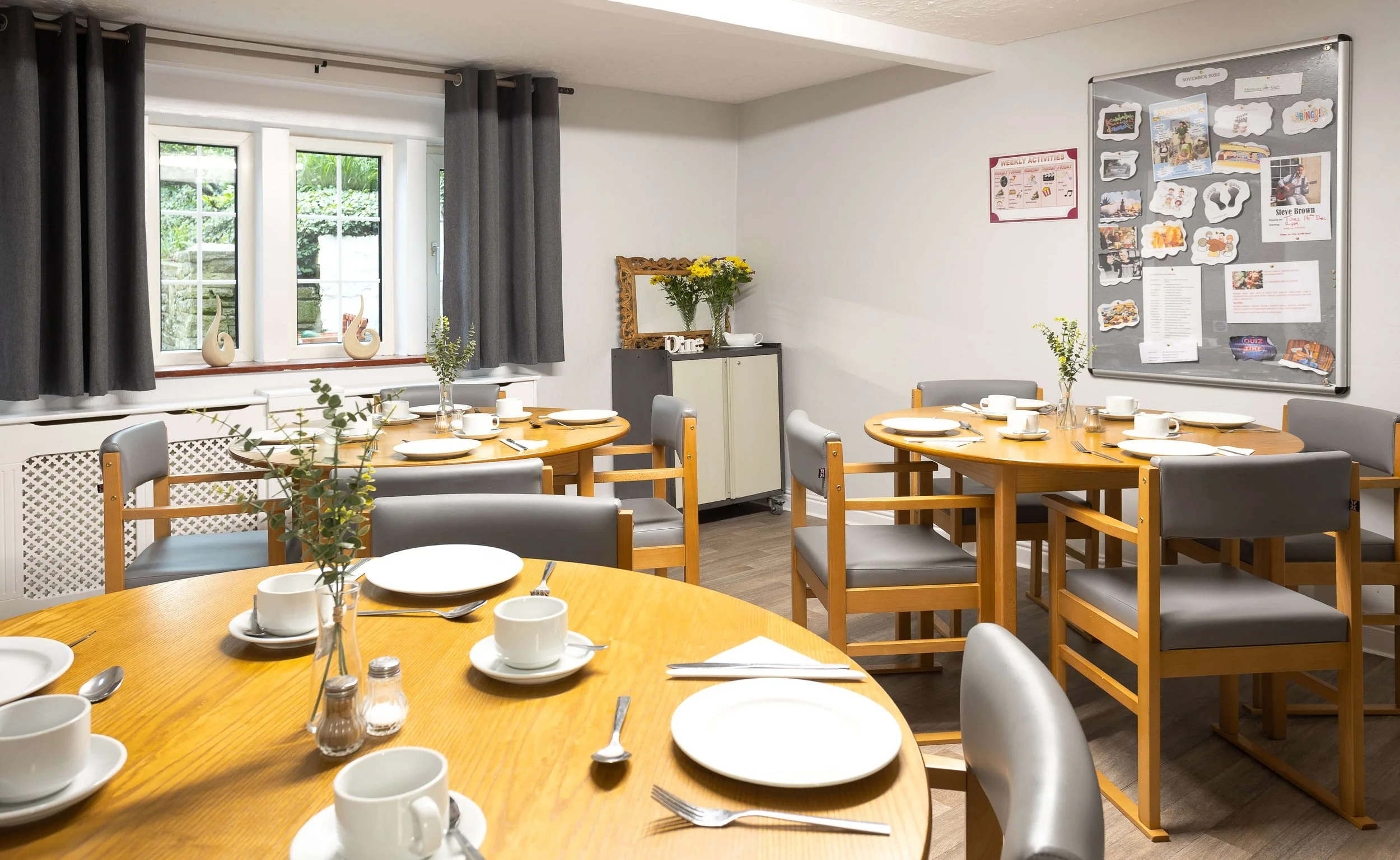 A dining room with three round wooden tables set with white plates, cups, saucers, and utensils. There are gray and wooden chairs around the tables and a window with dark curtains letting in natural light. A small cabinet with a vase of yellow flower