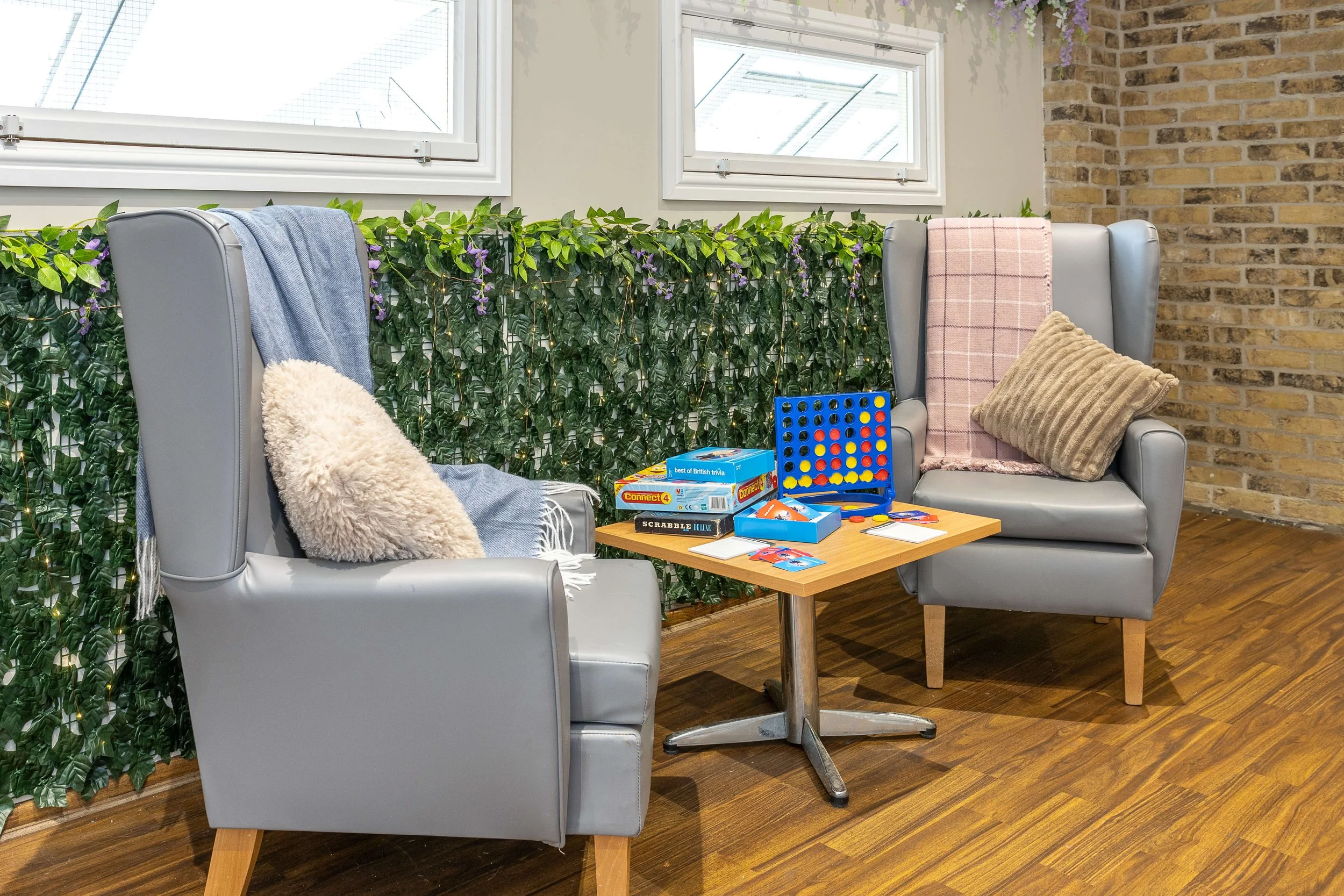 Two gray armchairs with cushions and throws, a small wooden table with board games and cards, and a decorative green leafy wall with string lights, windows, and a brick wall in the background.