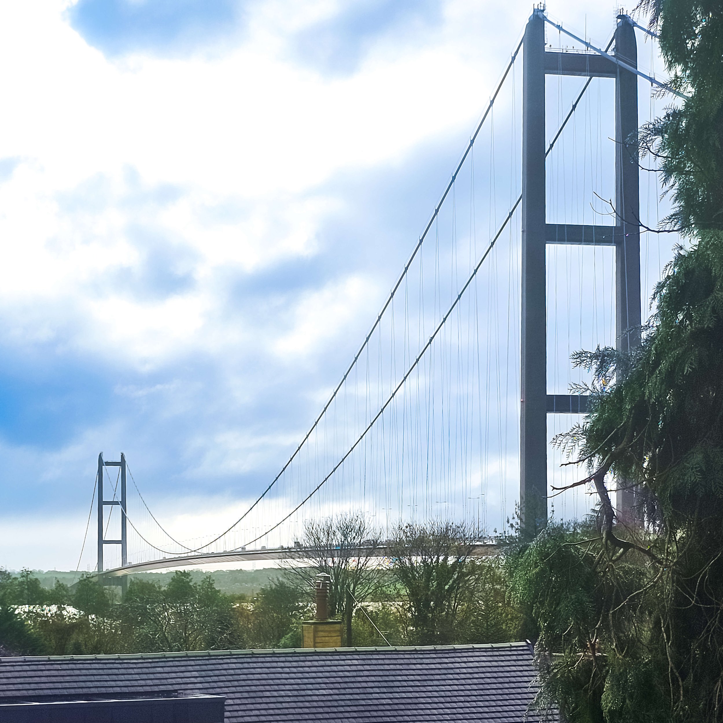 View from a bedroom window of the Humber Bridge across a landscape with trees and houses, under a cloudy blue sky.