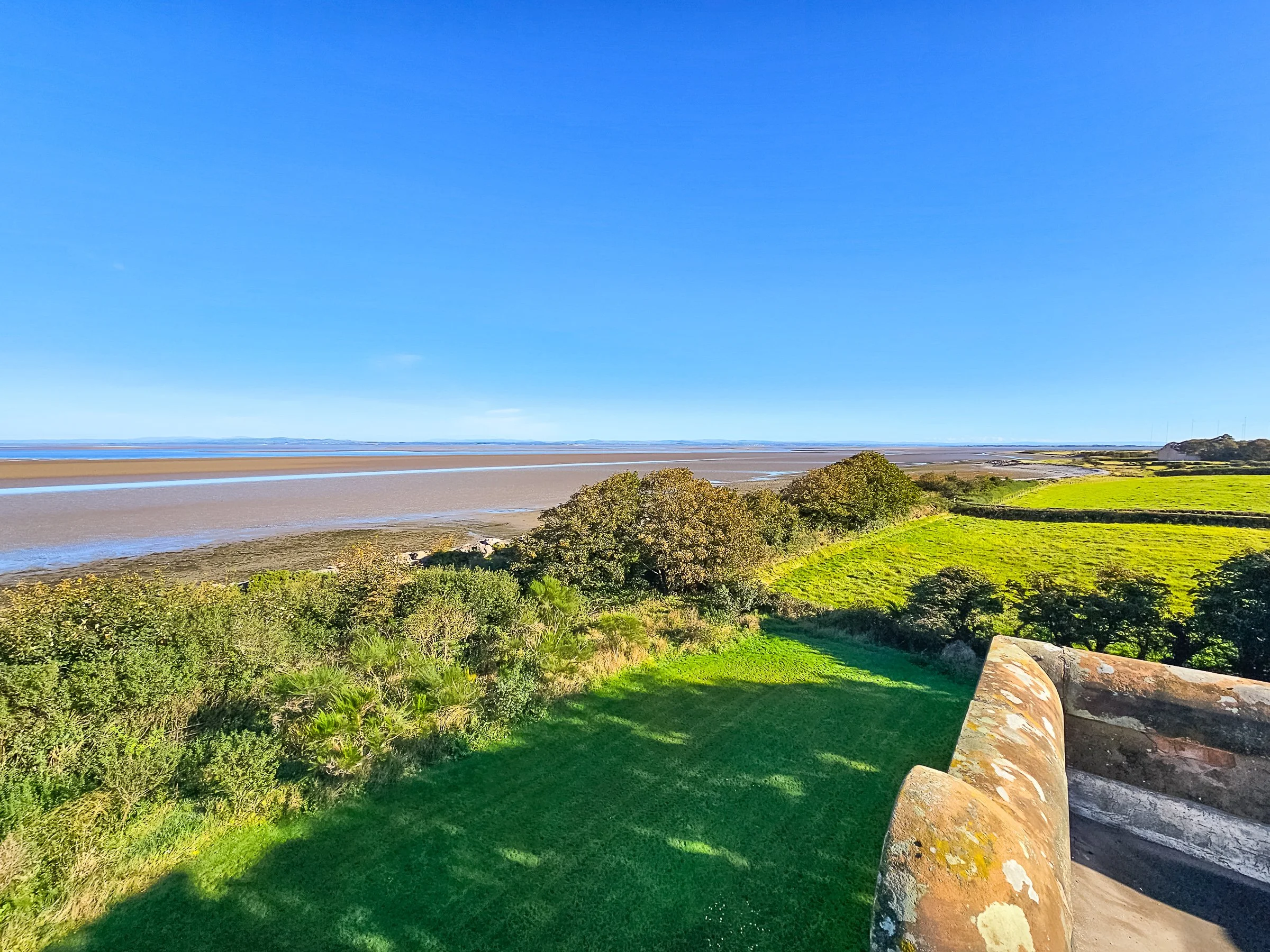 A coastal landscape with green fields and trees near a sandy beach under a clear blue sky.