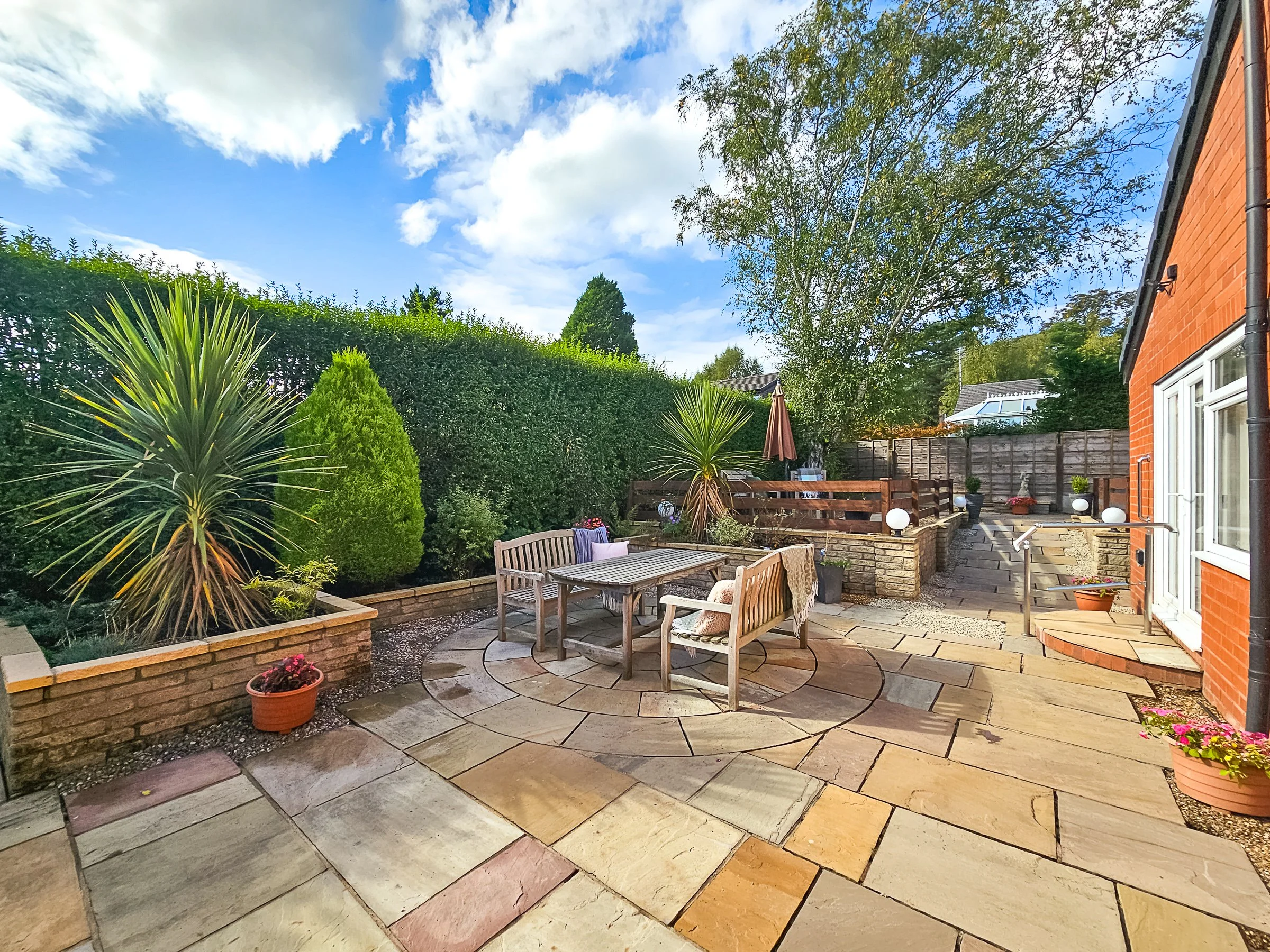 A backyard patio with stone paving, a wooden dining table with benches and chairs, lush green shrubs and trees, potted plants, and a wooden fence under a partly cloudy sky.