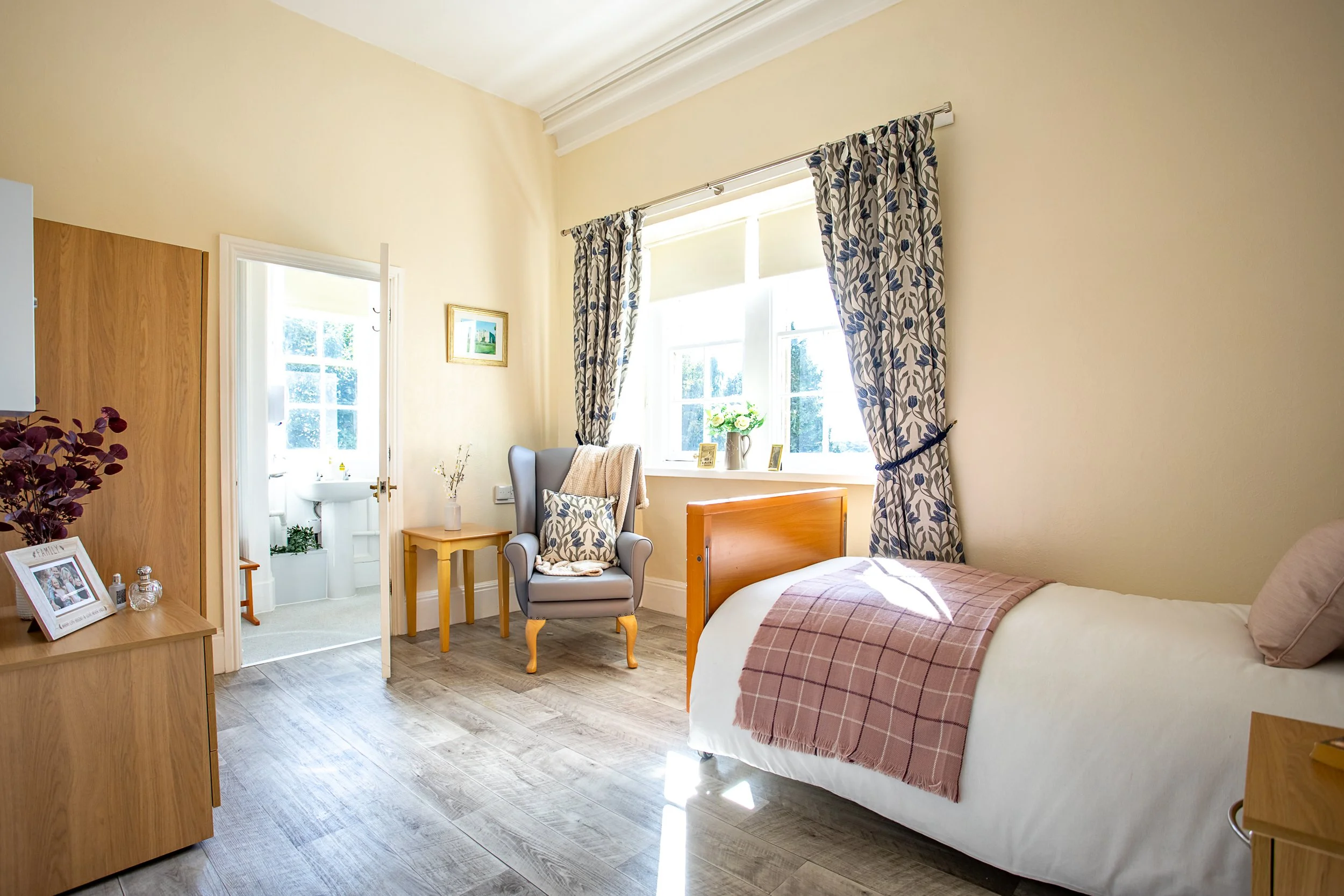 Bright bedroom with a bed, armchair, side tables, and a window with floral curtains, and an adjoining bathroom.