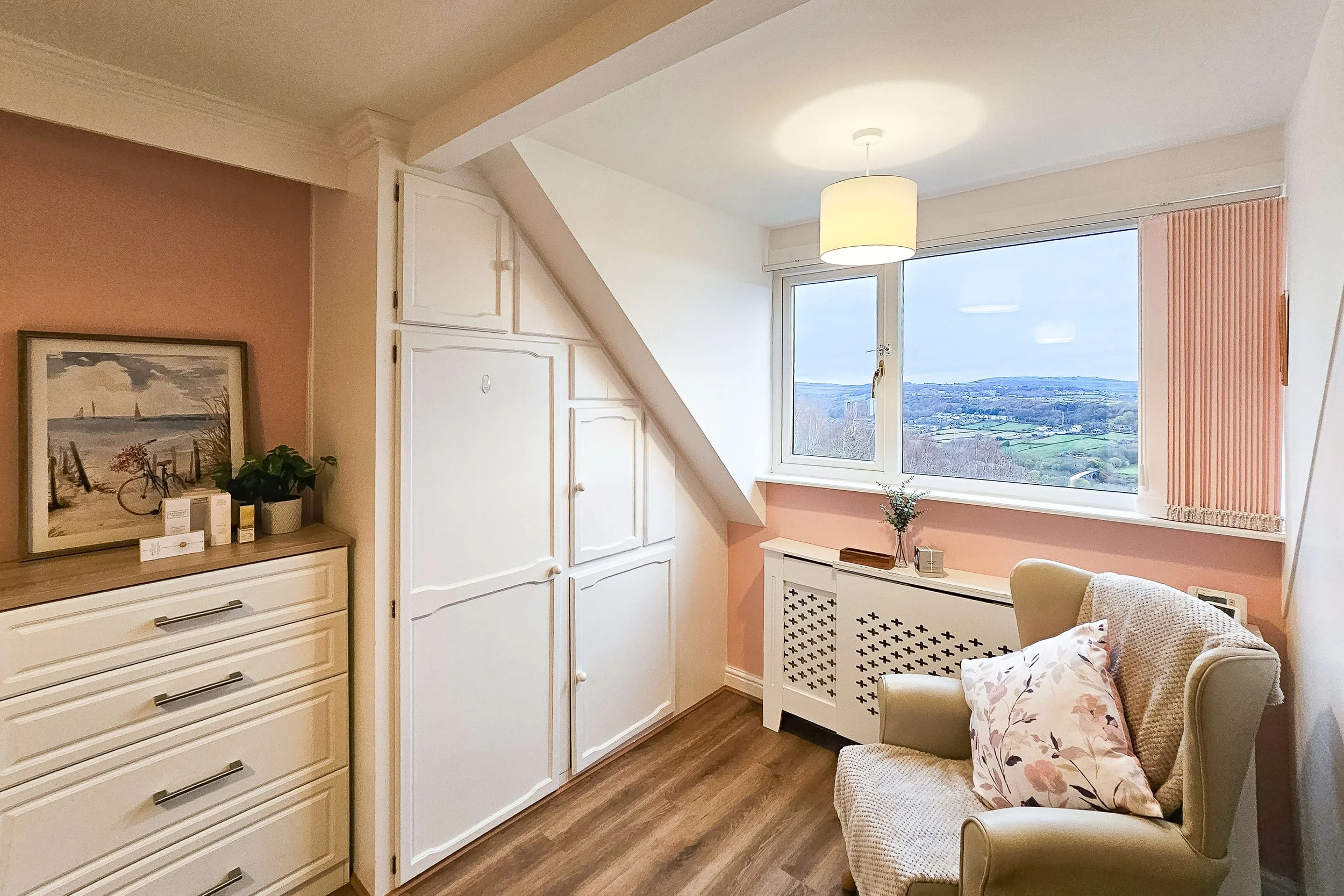 Cozy bedroom corner with a large window overlooking a landscape, an armchair with pillows, a dresser with decorative items, and built-in storage cabinets.