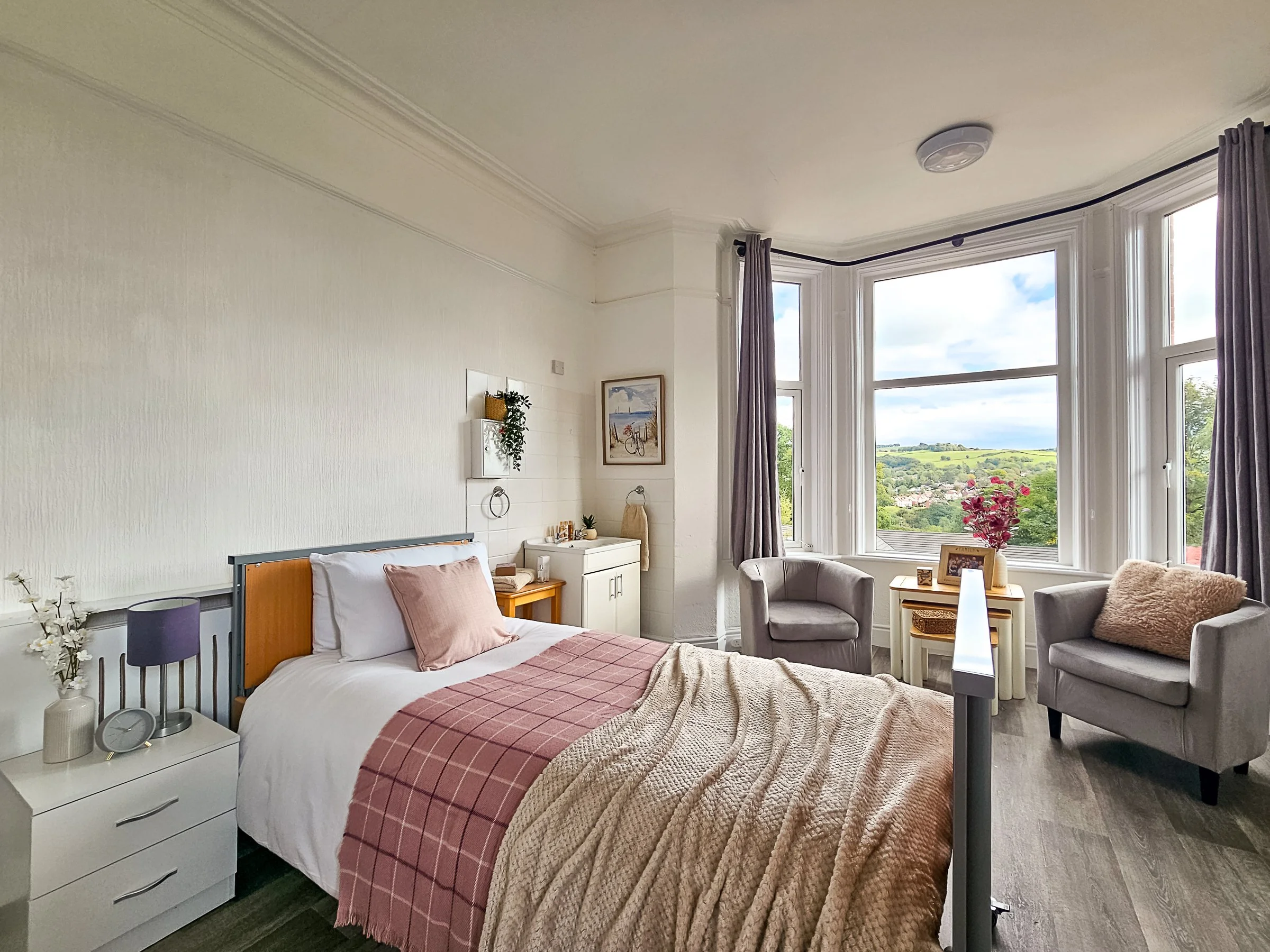 A cozy bedroom with a large bay window overlooking green hills. The bed has white linens, a pink pillow, and a beige throw blanket. There are two gray armchairs, a small wooden table with pink flowers, and various decorations.