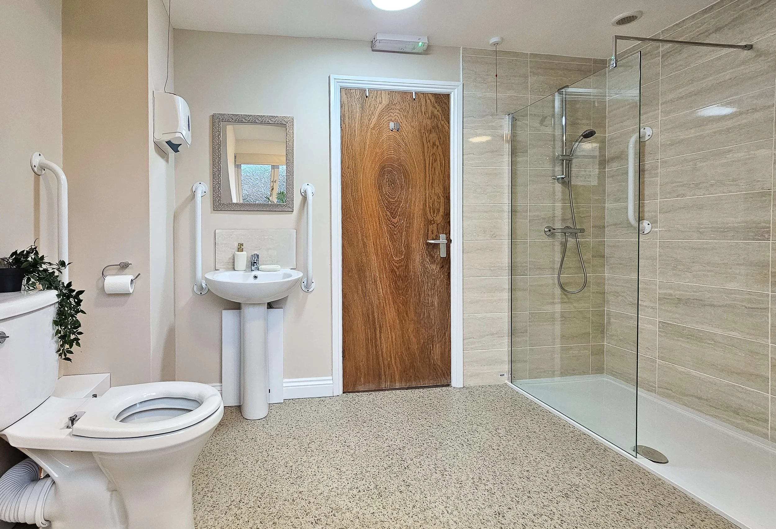A bathroom with a toilet, a small sink with safety bars, a mirror, a soap dispenser, and a shower area with a glass partition and a handheld showerhead, beige tiled walls, and a wooden door.