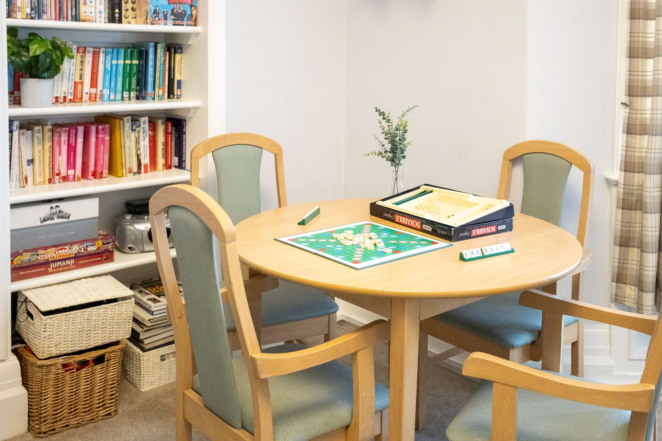 A round wooden dining table with four chairs, set up for a game of Scrabble, with game pieces on a board. There is a box of Scrabble game and a rack of letter tiles on the table. In the background, a shelf with books and a basket are visible, along w