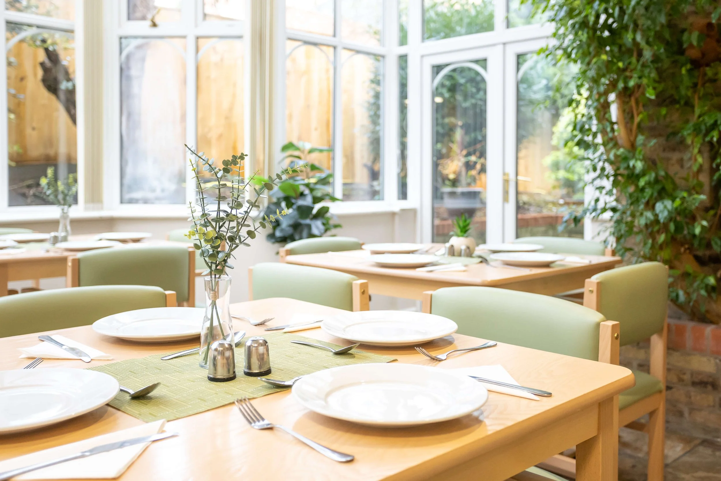 Sunlit dining area with wooden tables set with white plates, silverware, green placemats, and a vase with eucalyptus leaves, surrounded by green chairs and large windows with a garden view.
