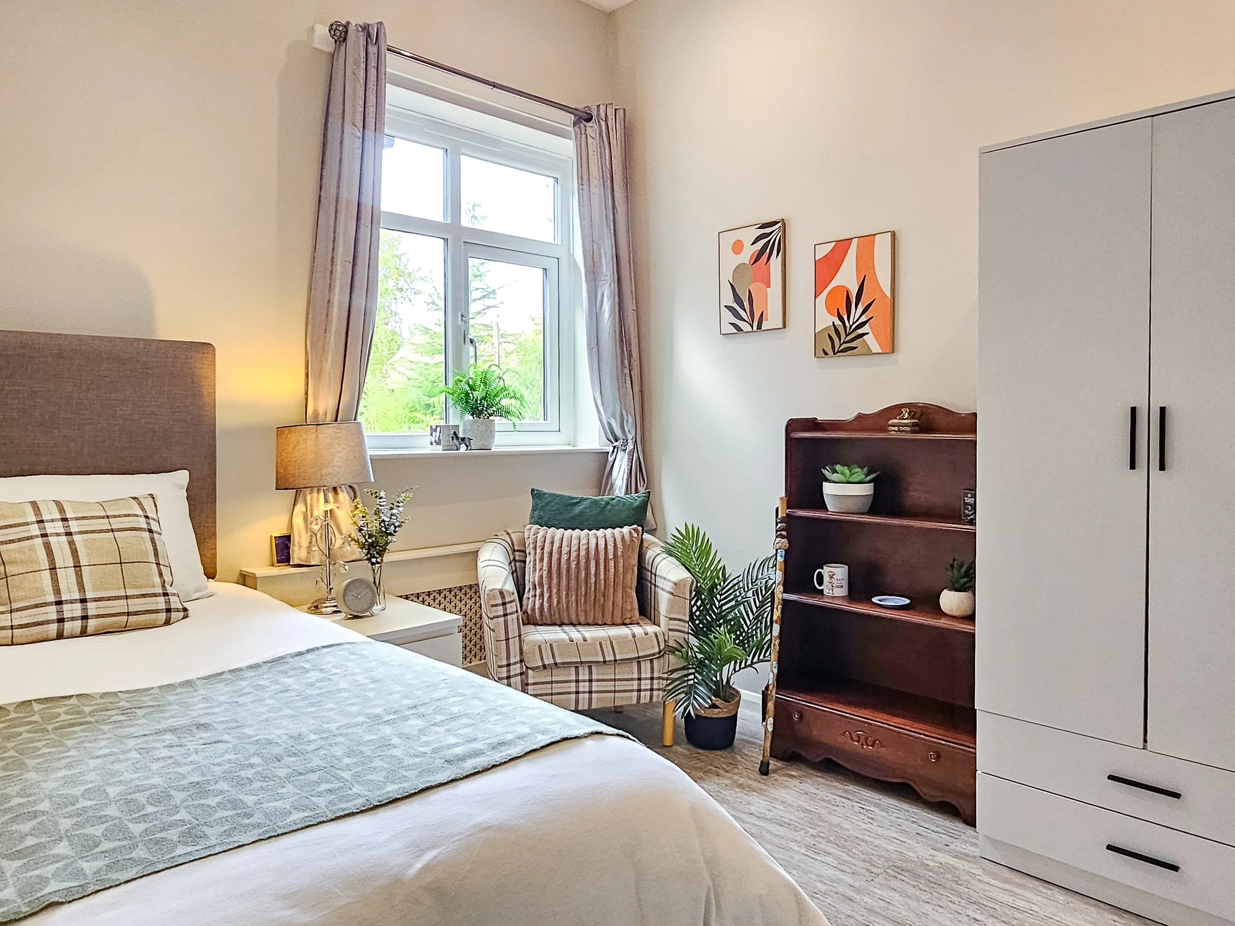 Cozy bedroom with a bed, bedside table with a lamp, armchair with cushions near window, framed abstract artwork on wall, potted plants, white wardrobe, and cream-colored walls.