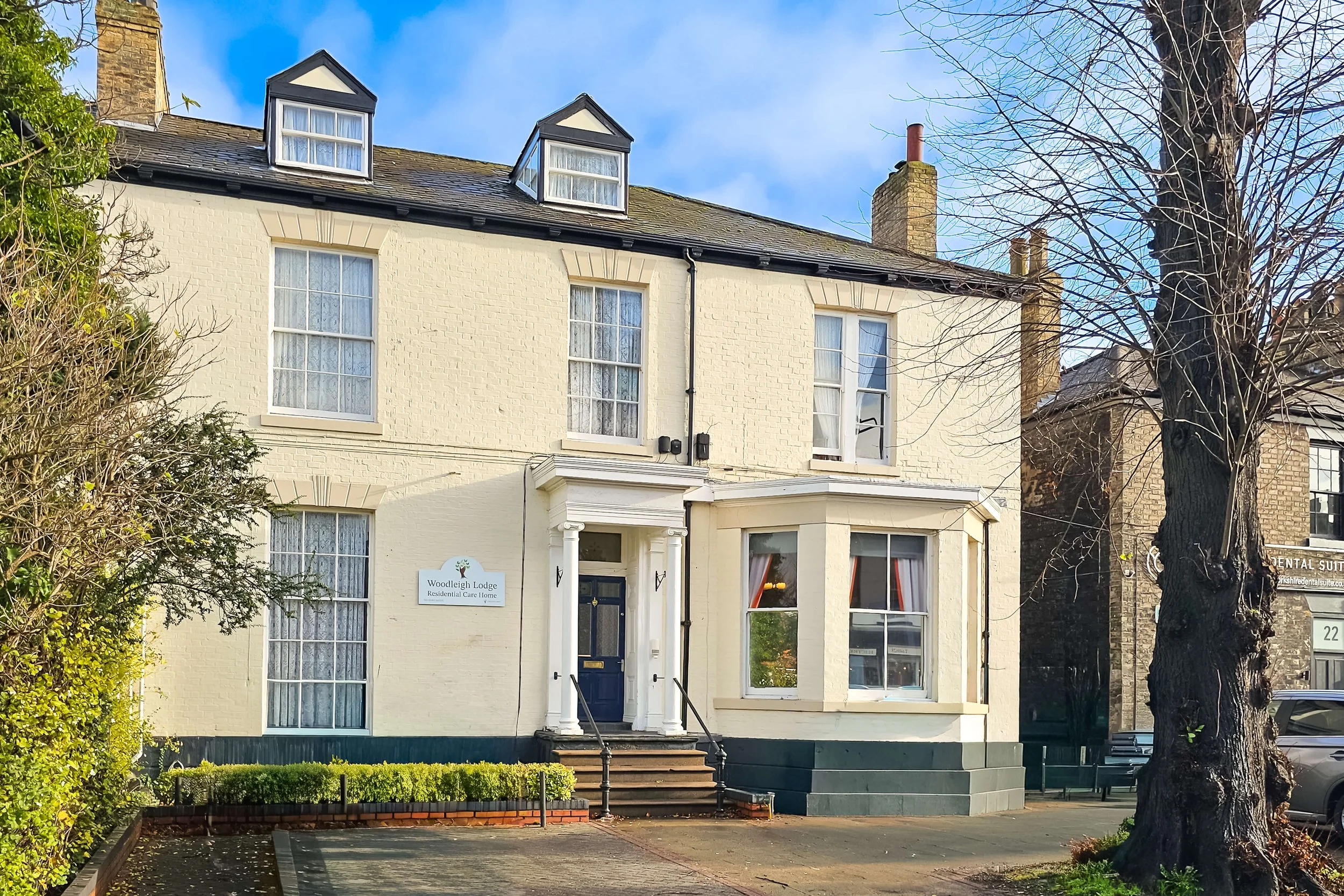 A three-story cream-colored building with a dark blue front door, steps, and a small set of railings. It has five windows visible on the front, with some with lace curtains. A sign reads 'Woodleigh Lodge Residential Care Home' on the left side of the entrance. There is a large tree on the right side and bushes on the left. The sky is blue with some clouds.