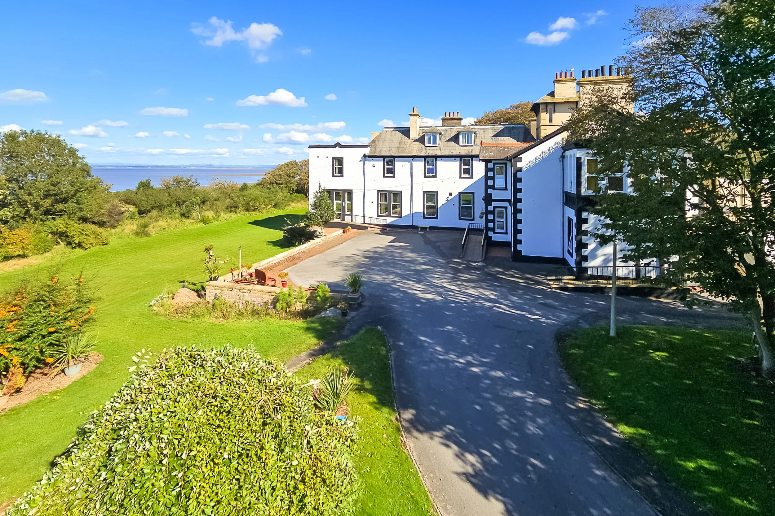 A large white care home with black trim and multiple chimneys, surrounded by green lawn and trees, under a blue sky with scattered clouds.