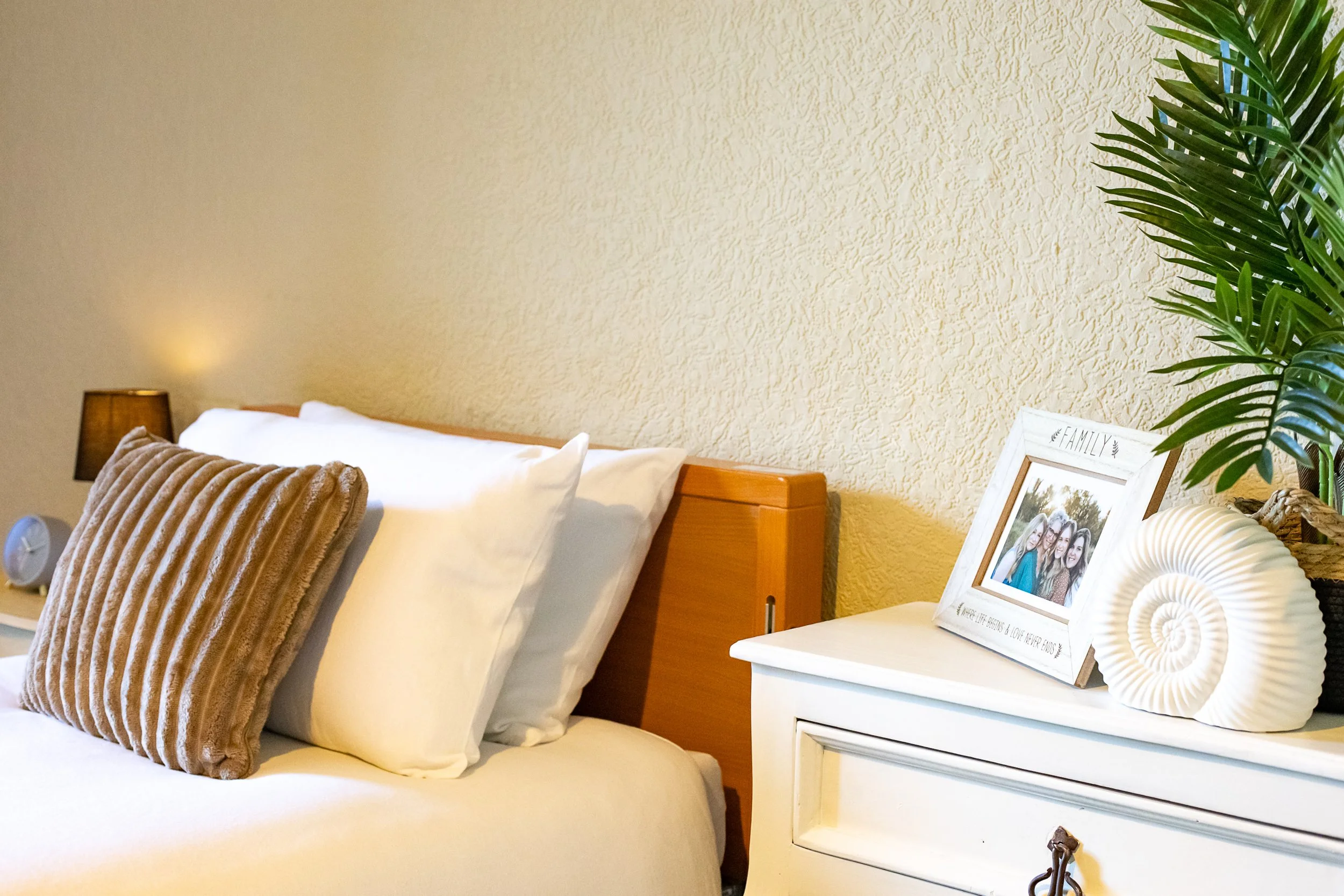 A neatly made bed with white pillows and a brown velvet decorative throw pillow. Next to the bed is a white nightstand with a family photo in a white frame, a large white seashell, and a green potted plant. A small lamp and alarm clock are on the nig
