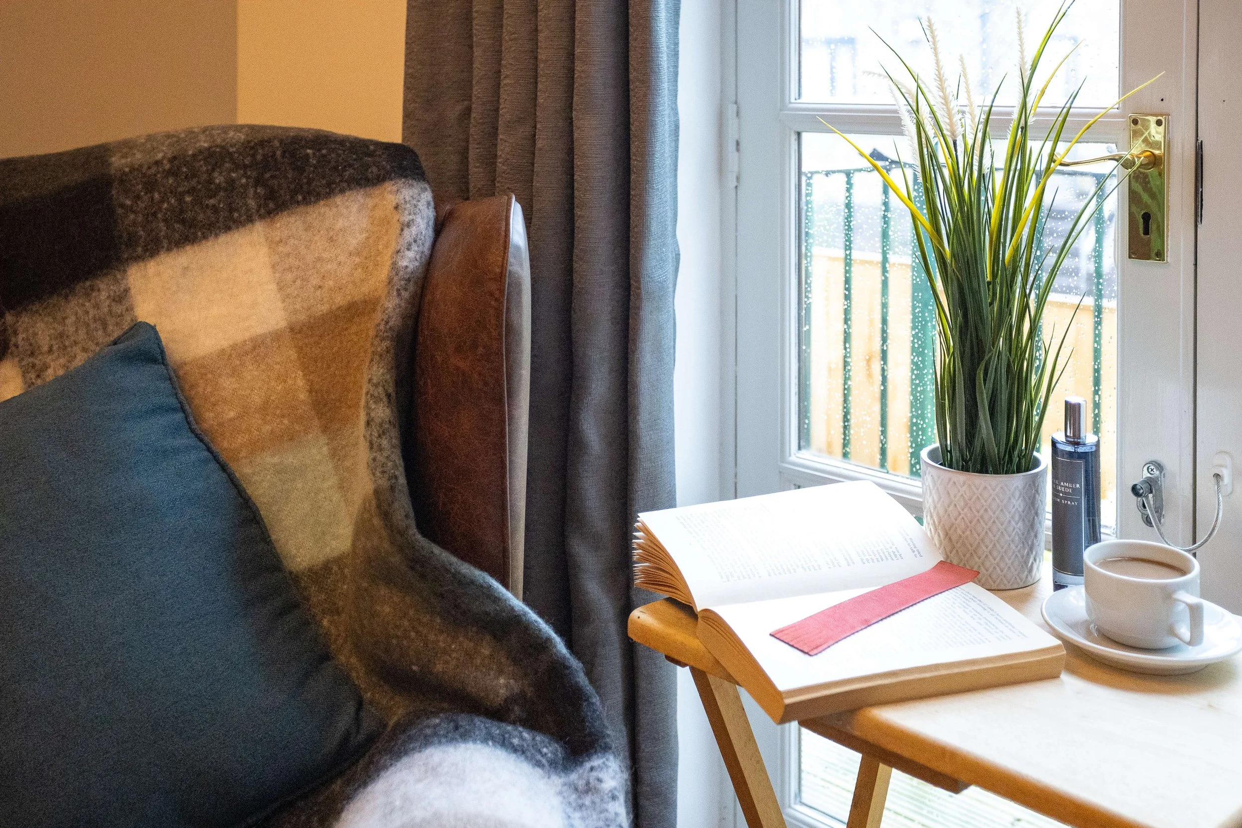 A cozy bedroom corner with an armchair, a side table with an open book, a potted plant, a cup of coffee, and a window with rain droplets and a balcony.