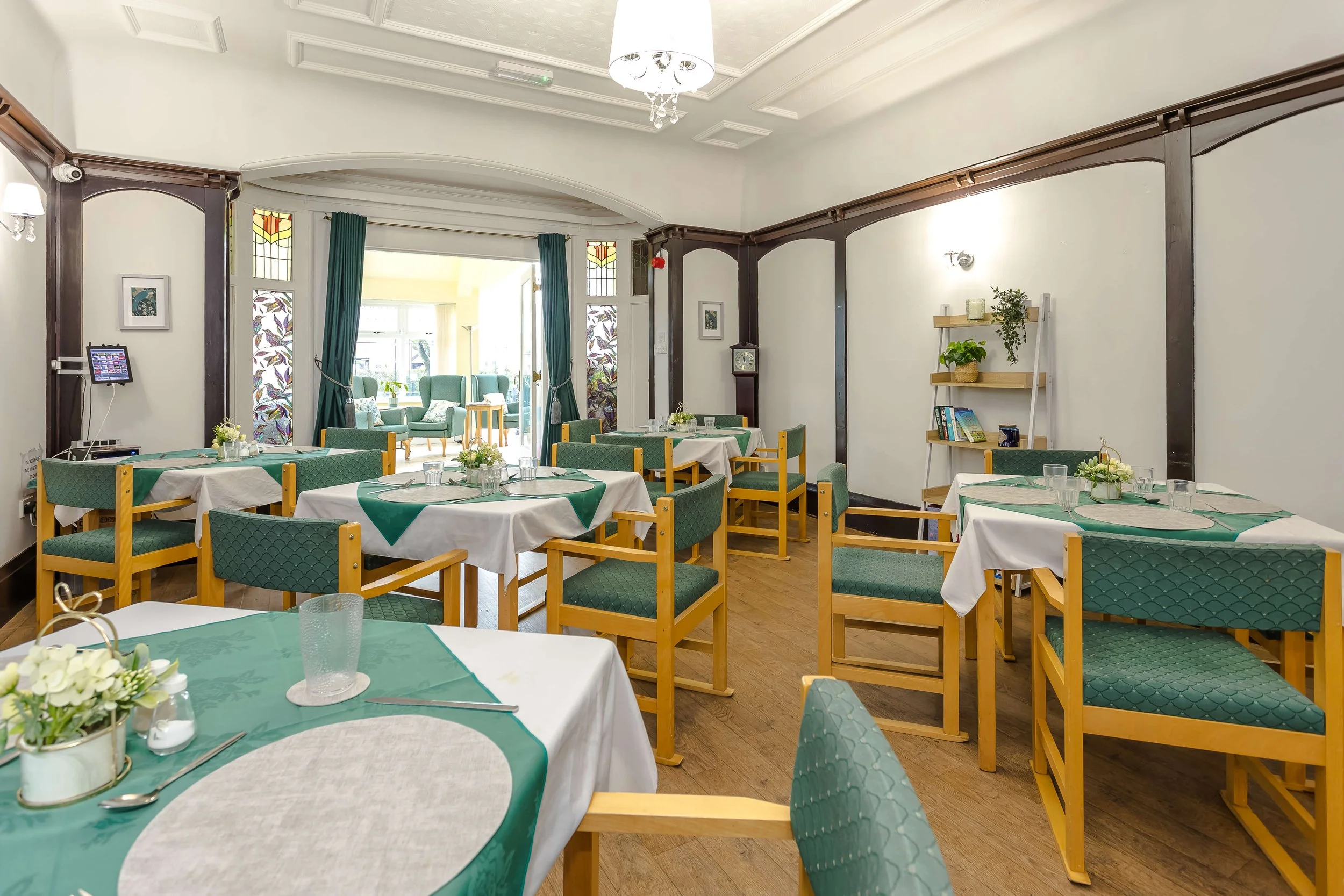 Dining room with tables set for a meal, decorated with green and white tablecloths, and chairs with green cushioned seats, large window in the background, and a bookshelf on the right side.