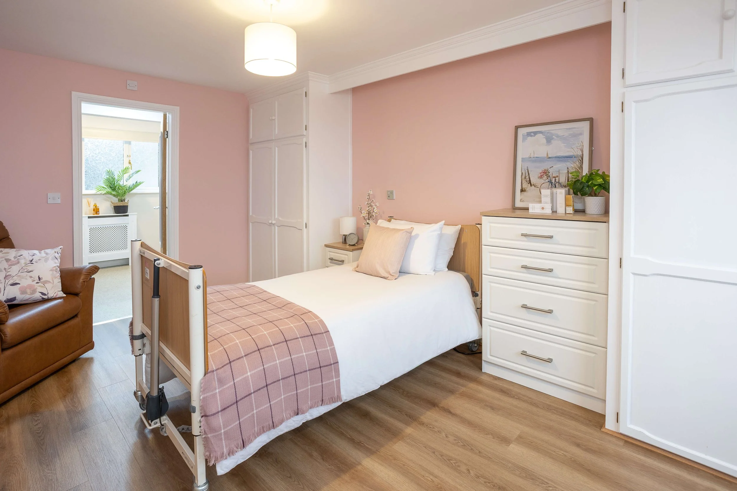 A bedroom with a pink accent wall, white bed with a beige throw blanket, and a hospital bed side table. There is a brown leather couch with floral pillows on the left, a drawer chest with artwork and a plant on top on the right, and a small nightstan