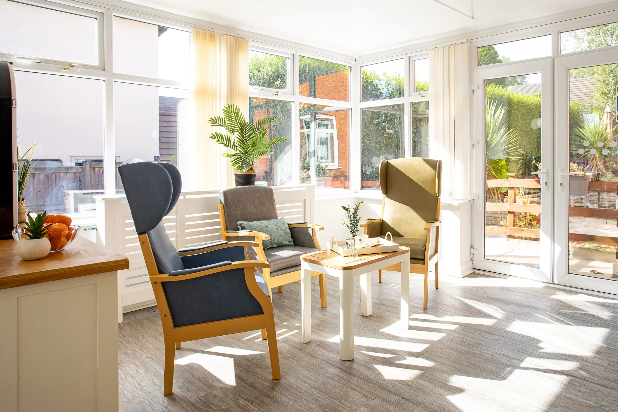 Bright sunroom with large windows, two armchairs, a small table with a plant, and outdoor plants visible through the windows.