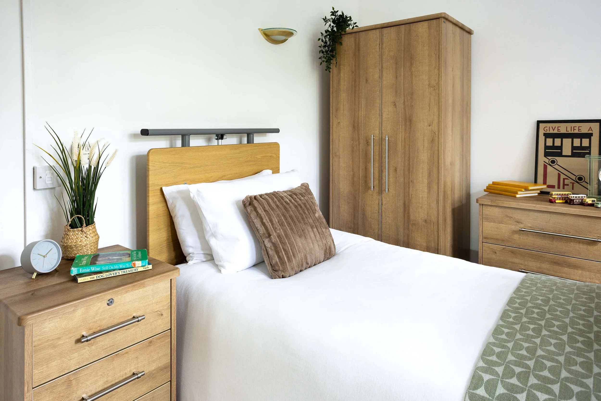 A tidy bedroom with a bed, beige pillows, wooden furniture including two dressers and a wardrobe, a green plant, books, a clock, and decorative items.
