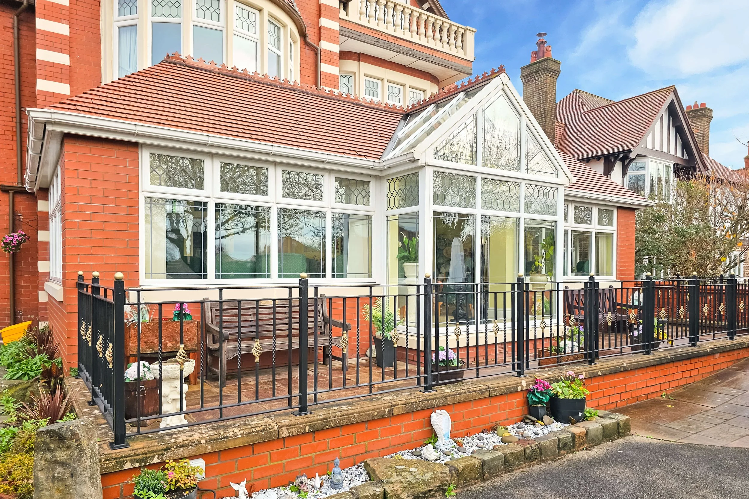 Front view of a brick house with a modern glass conservatory, surrounded by a black metal fence, potted plants, and garden ornaments.