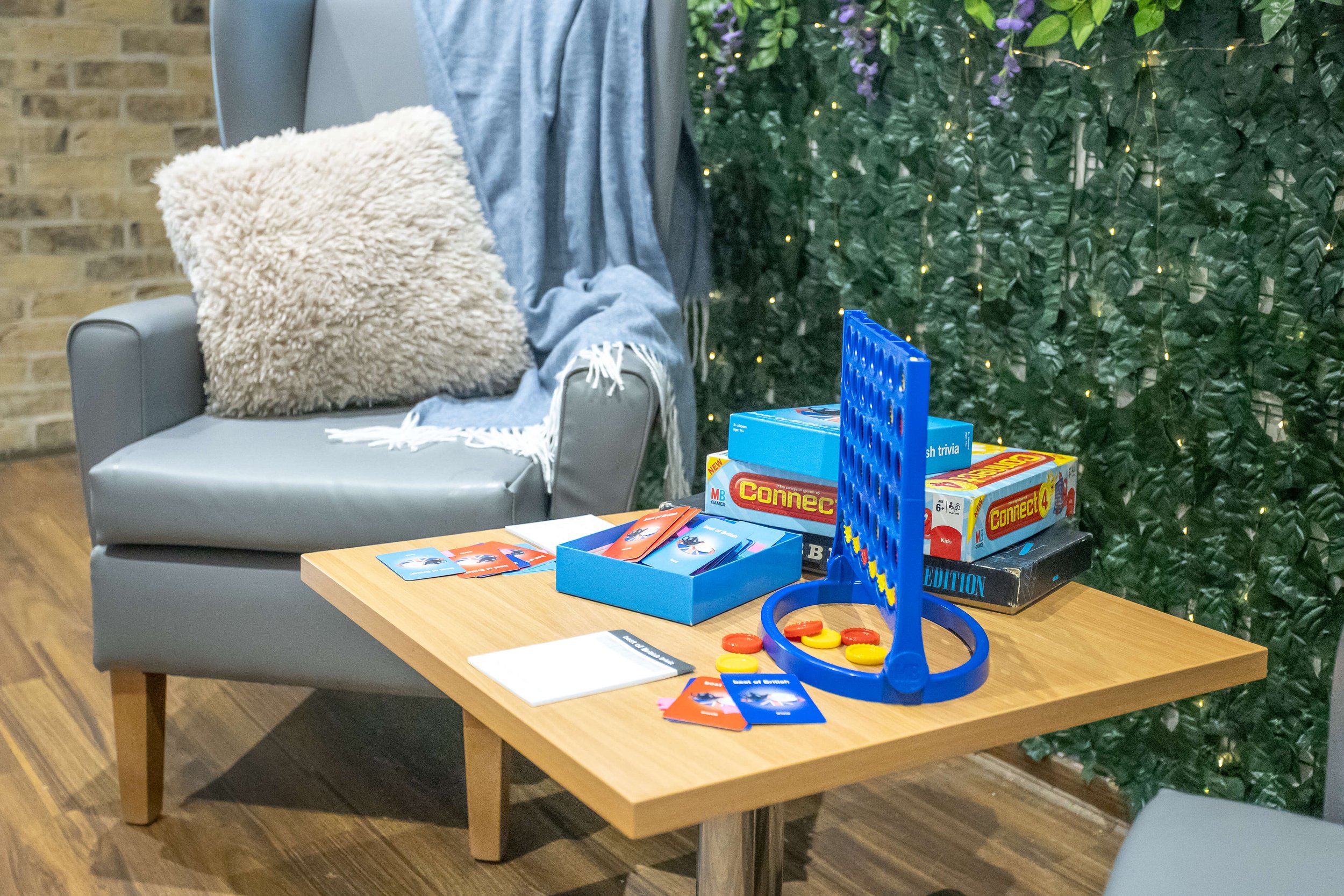 Table with board games, cards, and game pieces set up for playing. In the background, a cozy seating area with a gray armchair, a fluffy pillow, a blanket, and decorative greenery with fairy lights.