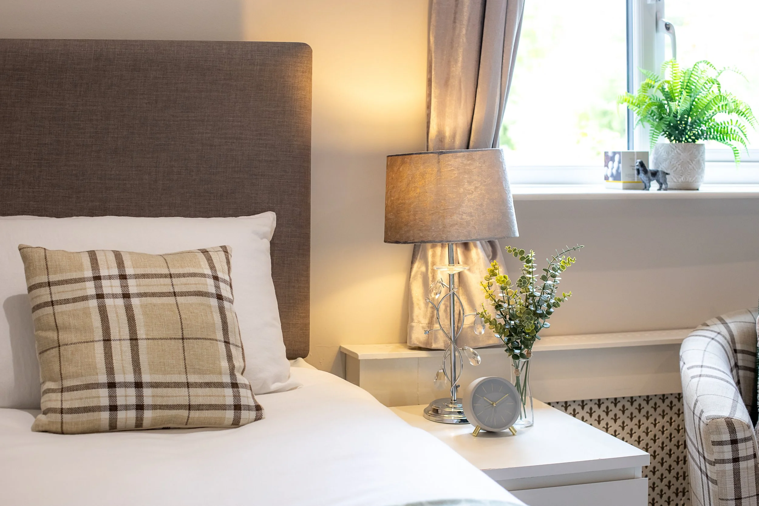 A bedroom with a bed that has white sheets and a beige plaid pillow. A nightstand with a lamp, a clock, and a vase of green plants. A window with beige curtains and decorative items on the sill.