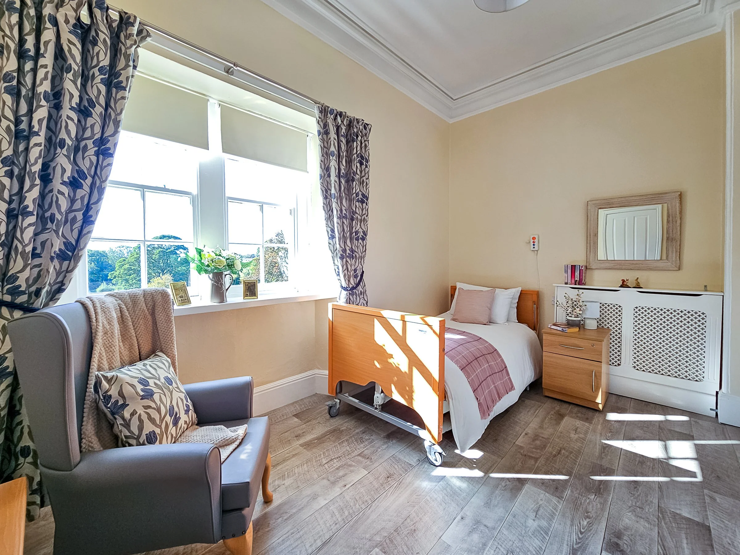 A cozy bedroom with a single bed near a large window, natural sunlight streaming in, and a grey armchair with a patterned pillow, with light-coloured walls and wooden flooring.