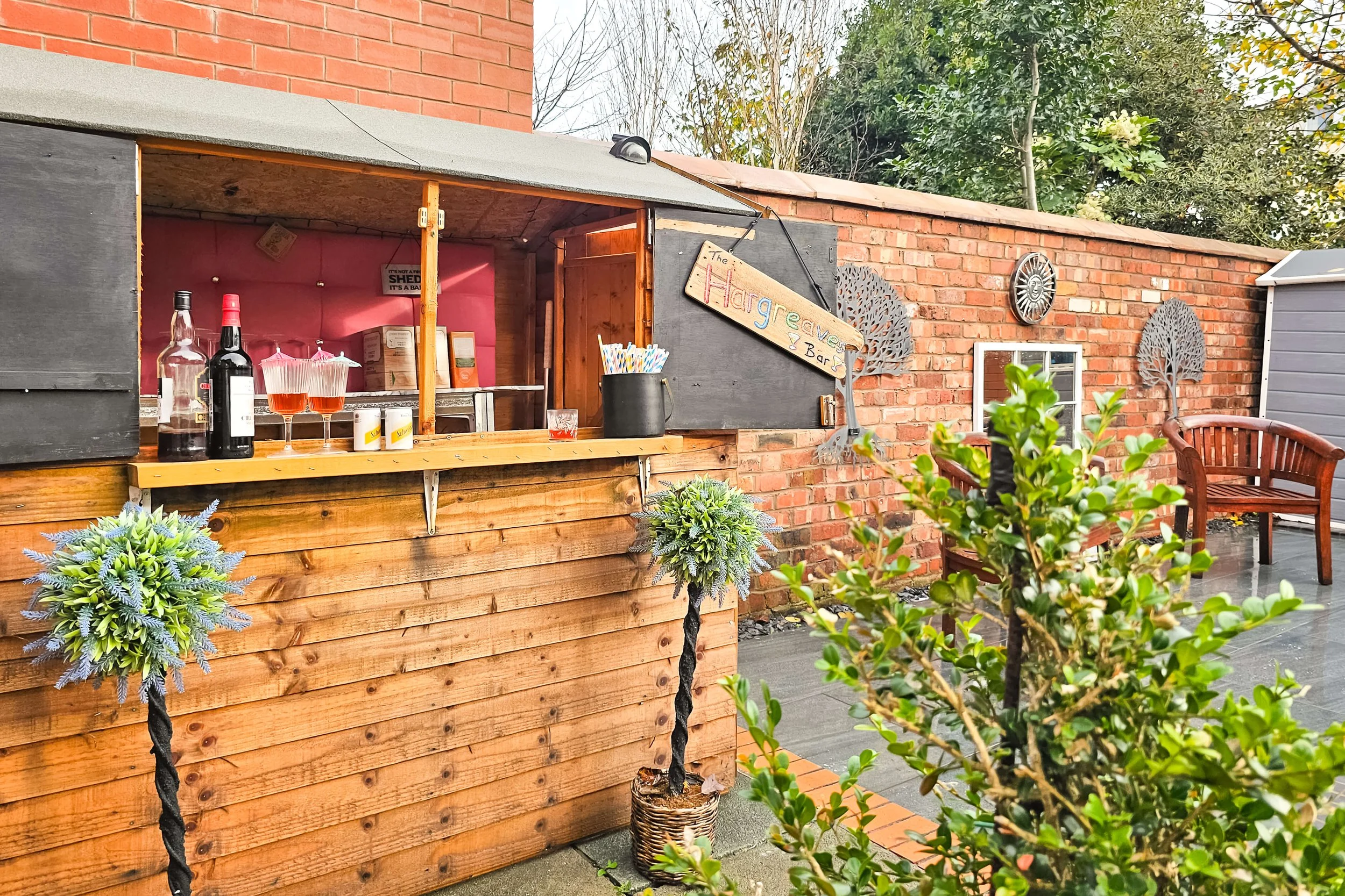 Outdoor bar with wooden counters, bottles of alcohol, glasses, and a pot with straws. There's a sign reading "The Hargreaves I Bar" and decorative metal trees on a brick wall with a sitting area and greenery.