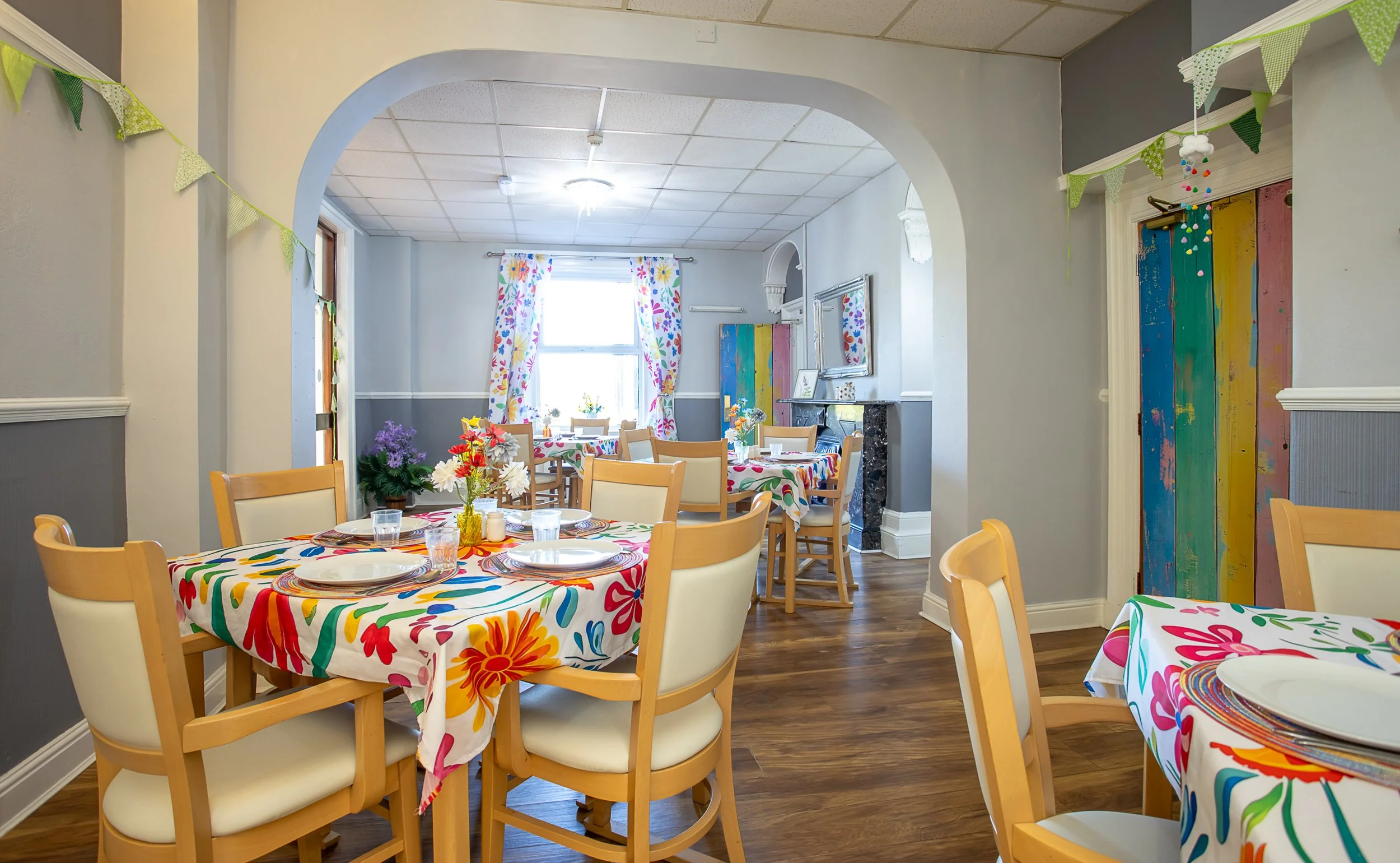 Colorful, festive dining room with round tables covered in floral tablecloths, set with plates and glasses. Bright curtains with a floral pattern, decorative bunting, and a multicolored wooden door add to the cheerful decor.