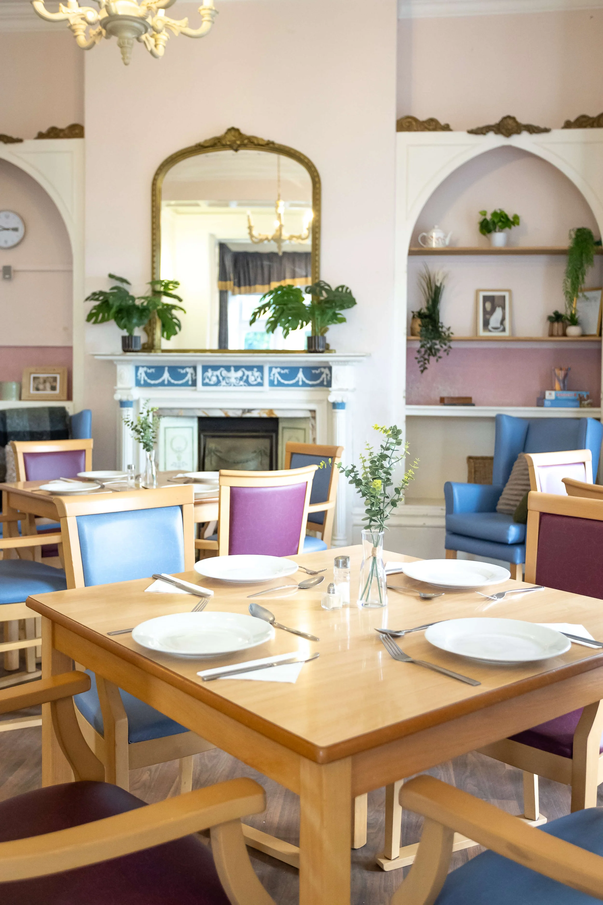 A cozy dining room with a wooden table set with white plates, silverware, glass vases with greenery, and a decorative fireplace with a mirror above it and potted plants. There are colorful chairs around the table and a blue armchair in the corner, wi