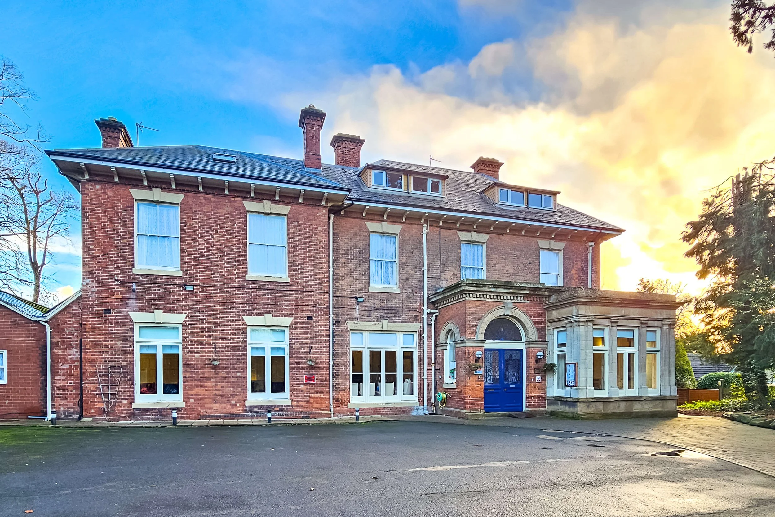 A large, three-story brick house with white window frames and a blue front door, set against a sky with a mix of blue and orange hues at sunset.