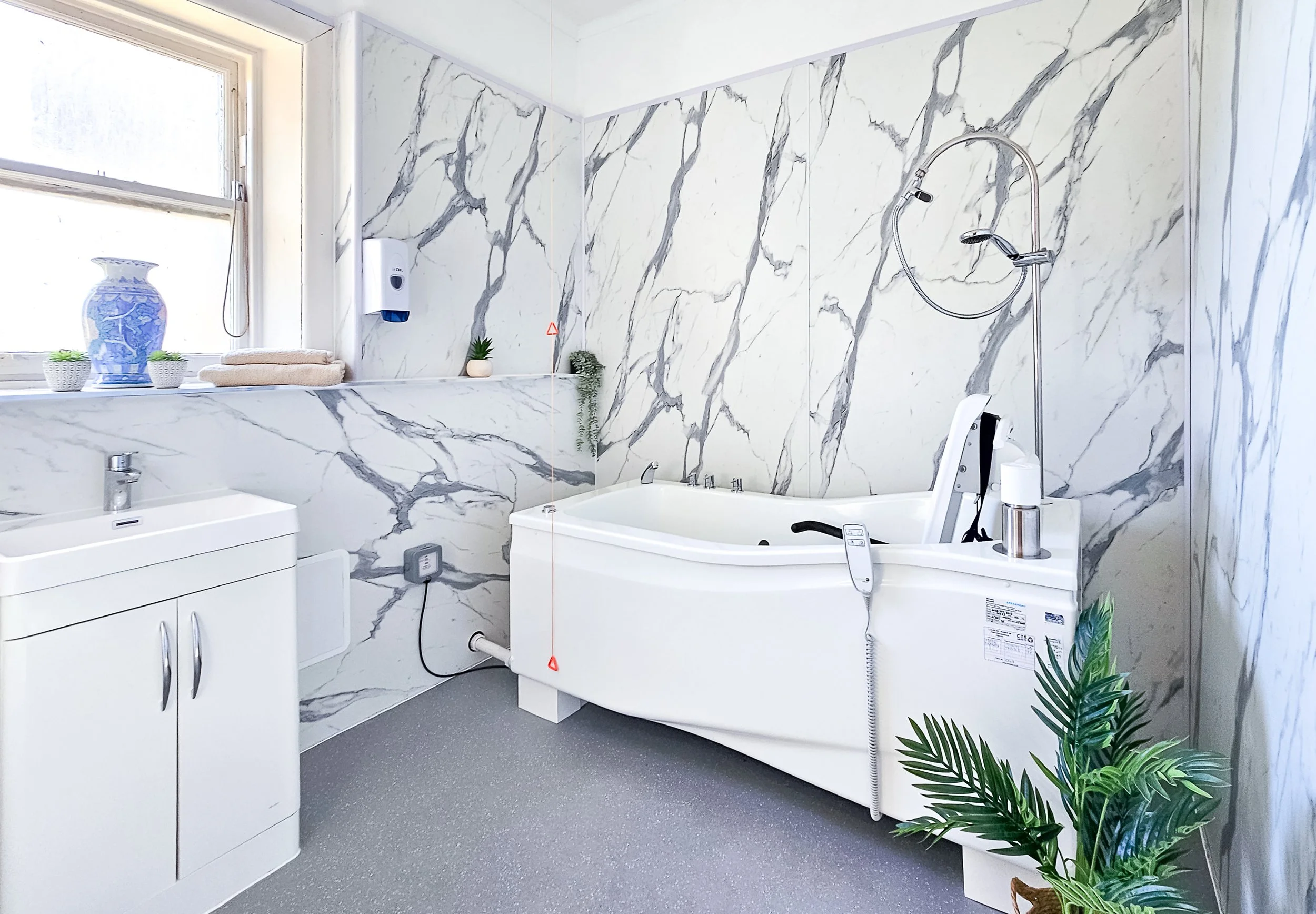 Modern bathroom with marble walls, a white sink cabinet, a window, small decorative plants, towels, a mounted soap saver, and a white bathtub with a handheld shower head, controller, and personal care items.