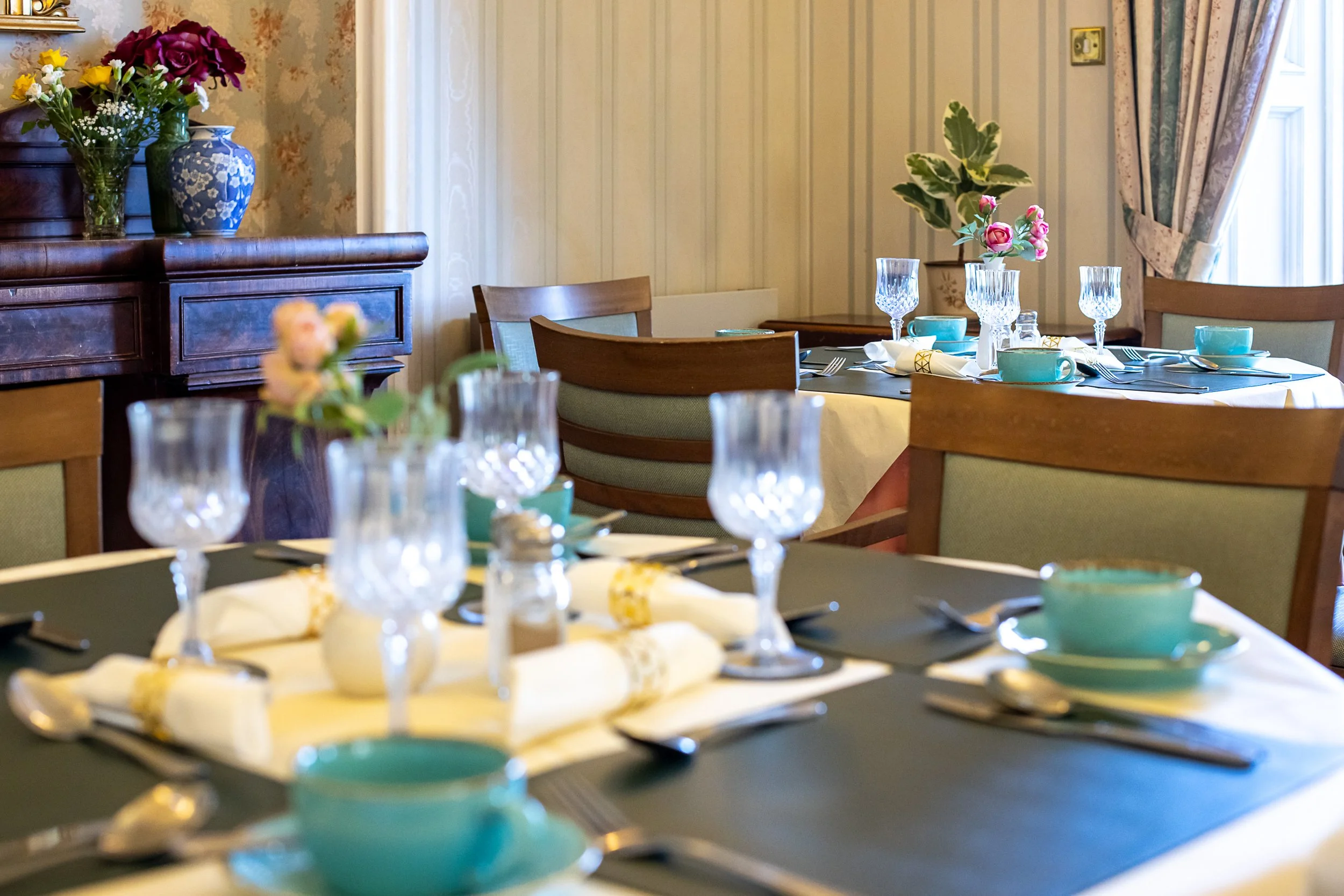Dining room with a table set for a meal, including teal cups, glasses, silverware, and a flower arrangement, with a wooden sideboard and floral curtains.