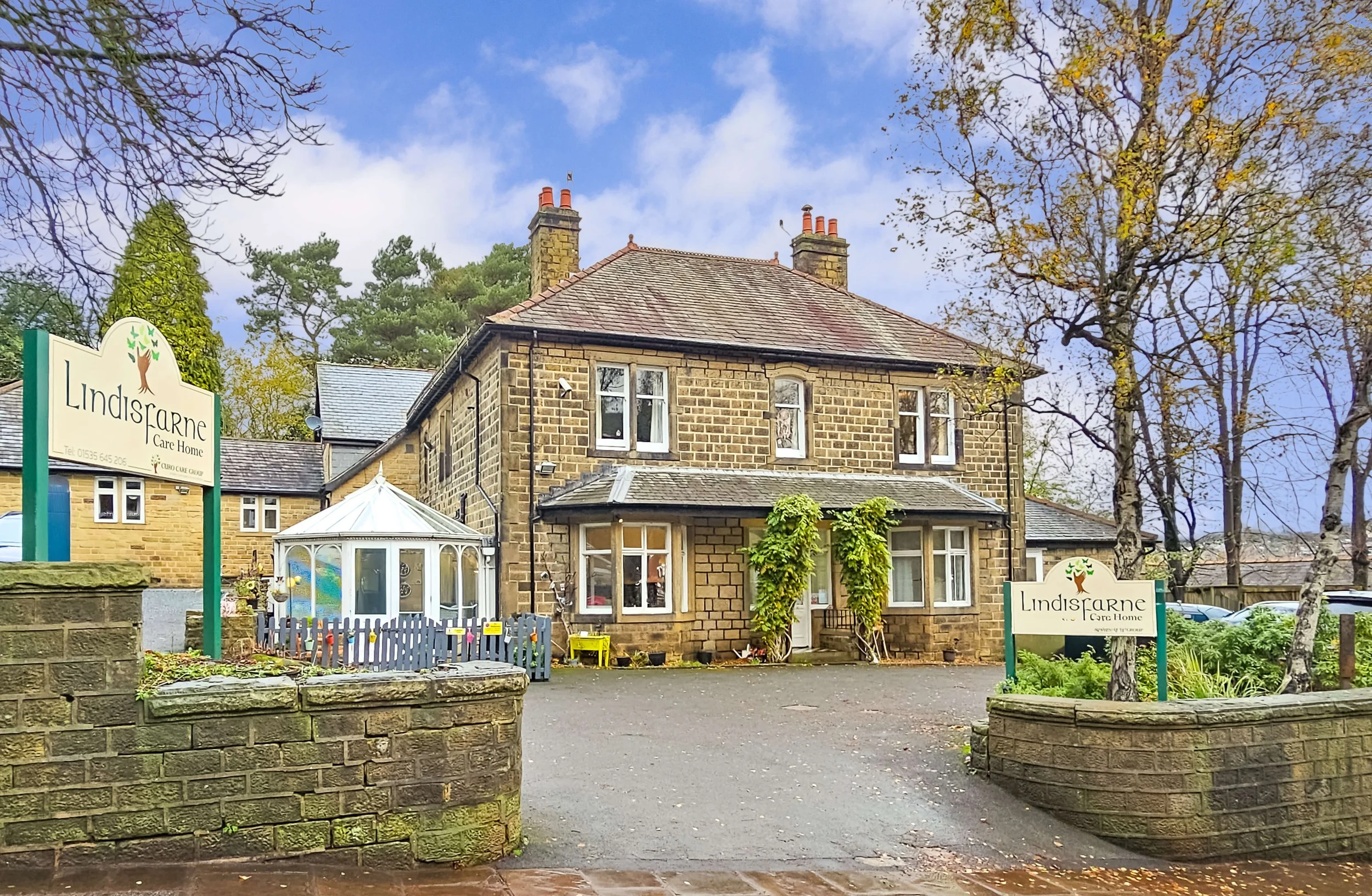 A stone care home building with a gabled roof, surrounded by trees with autumn leaves, and two signs that read 'Lindisarne Care Home' on a brick wall and a small garden area.