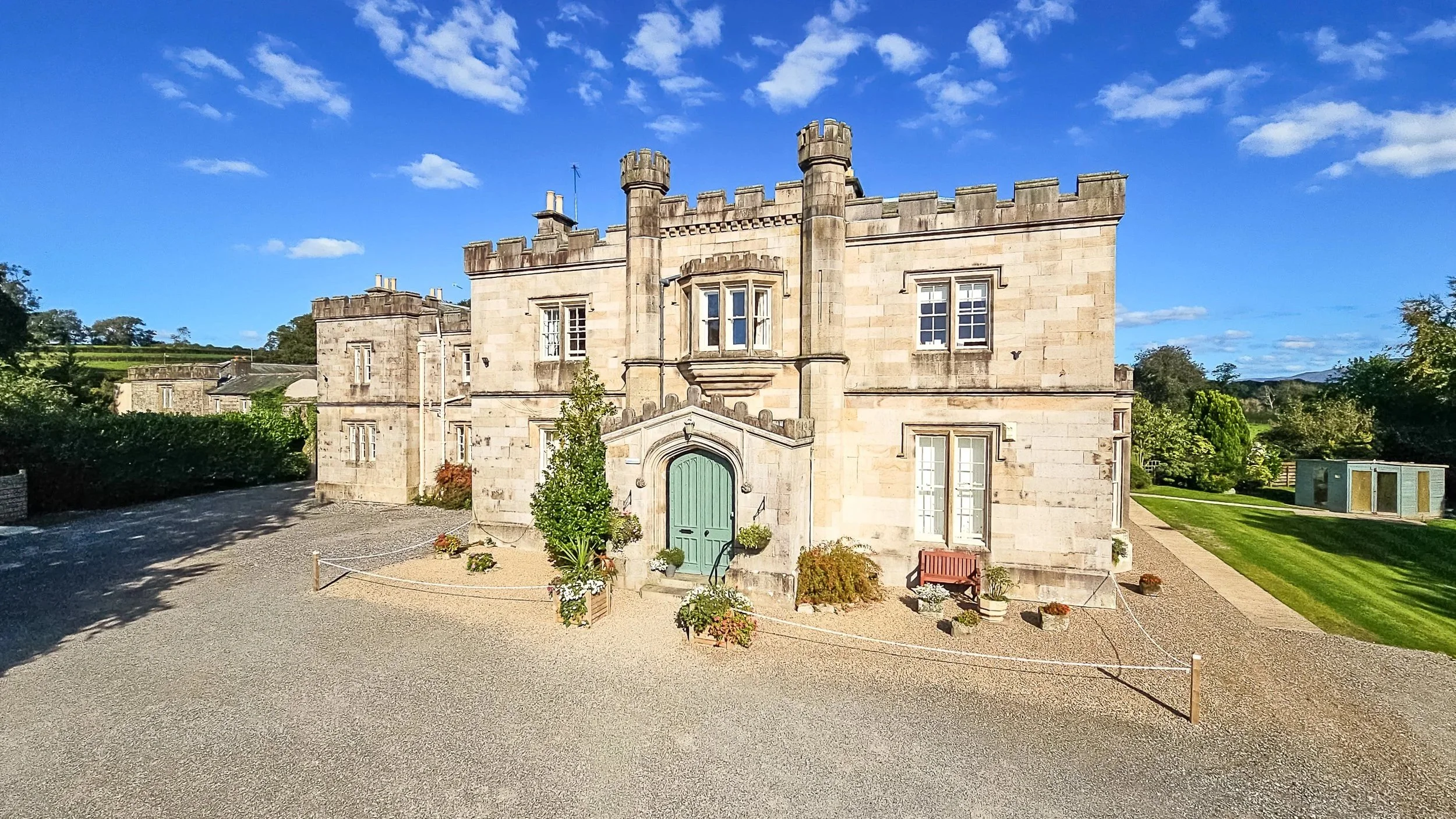 A stone castle-like building with battlements, multiple windows, and a centered arched doorway painted green. There are potted plants, shrubs, and a wooden bench outside the entrance. The building is set in a landscaped area with a gravel driveway and green lawns, under a bright blue sky with scattered clouds.